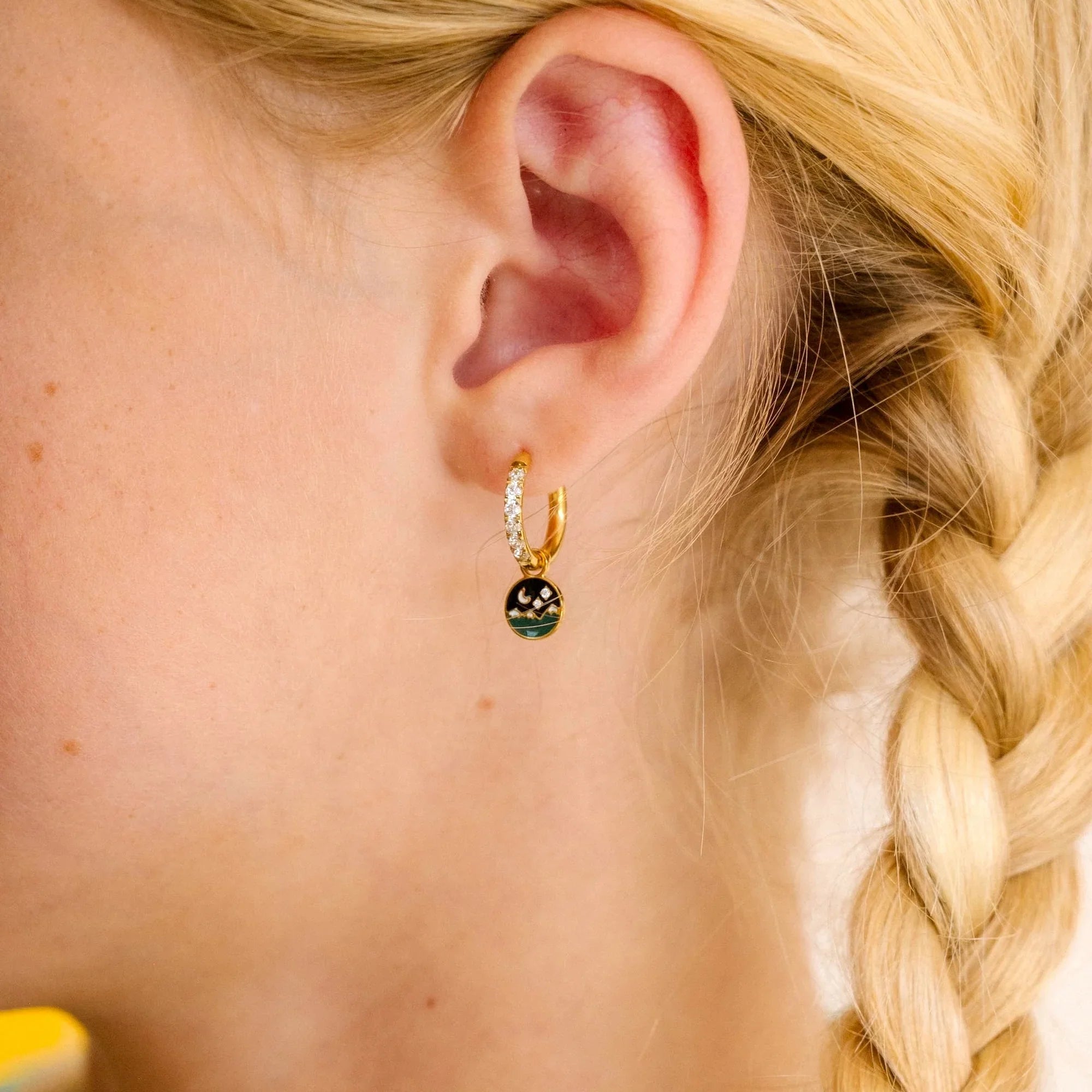 Close-up of an ear wearing a gold earring with a night sky scene made from enamel and clear gemstones, blonde hair in a braid.