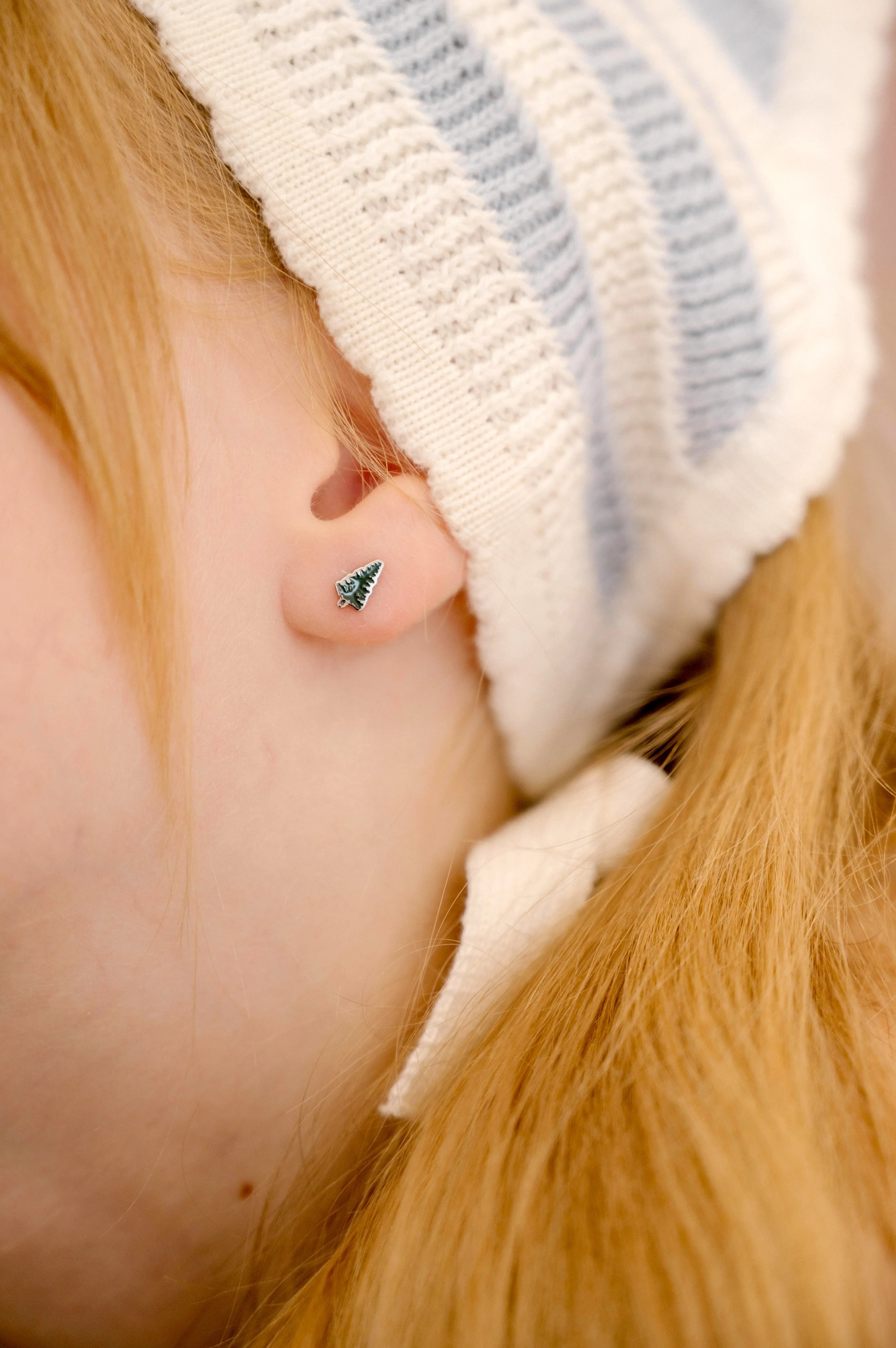Close-up of a child wearing a white knit hat with blue stripes, focusing on the ear with a green enamel pine tree-shaped earring.