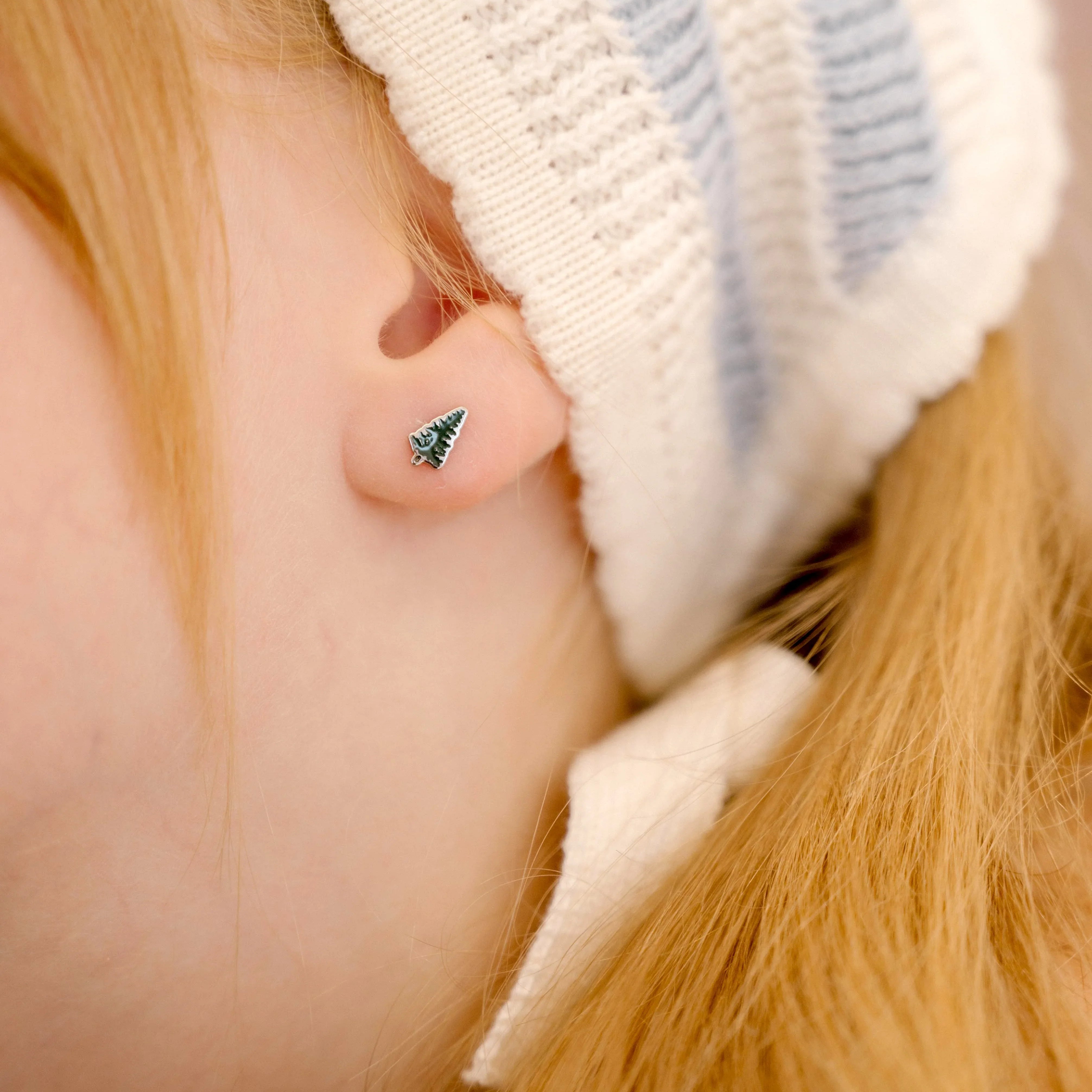 Close-up of a child wearing a white knit hat with blue stripes, focusing on the ear with a green enamel pine tree-shaped earring.