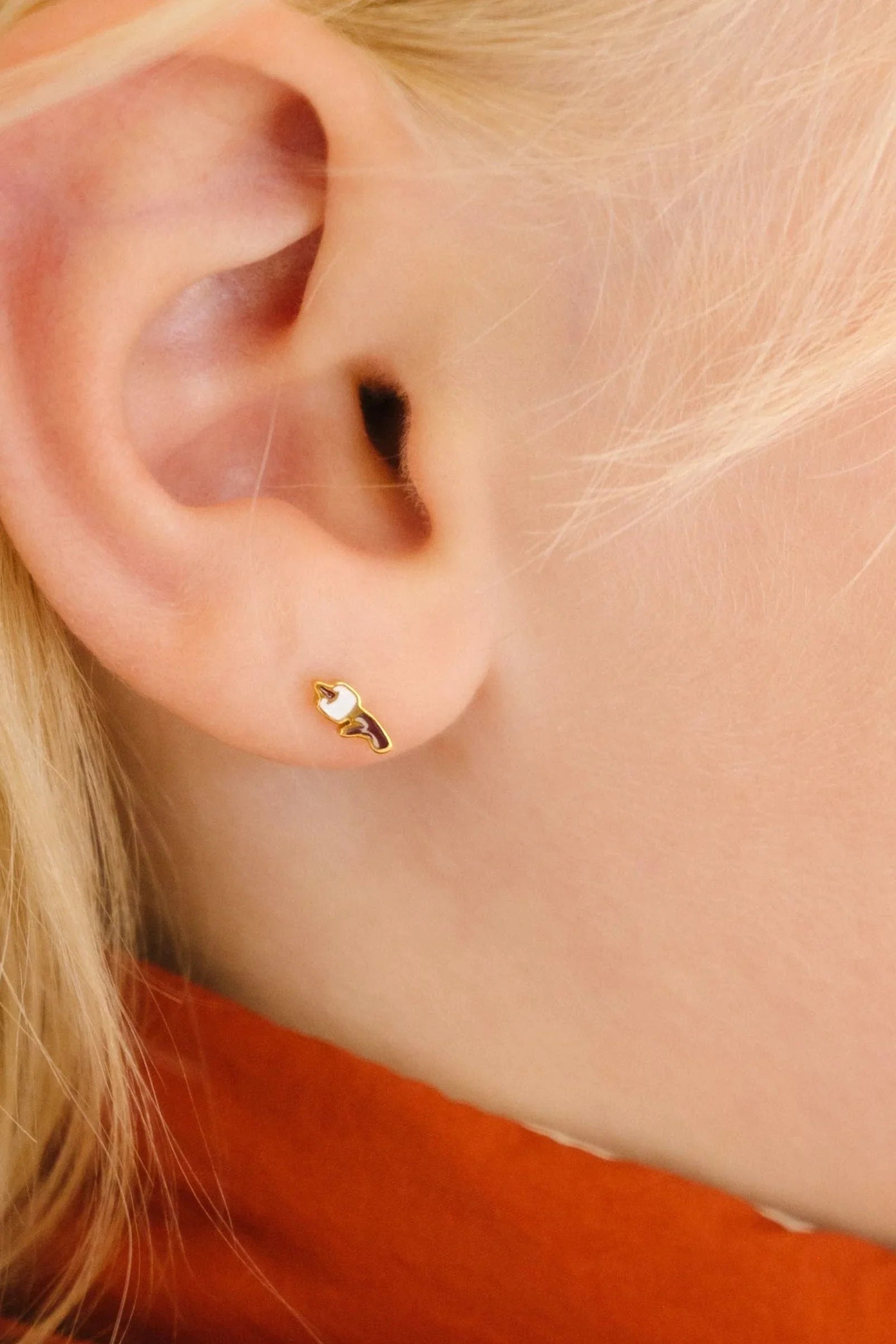 Close up of a blond person's ear with a gold-colored stud earring with a brown and white enamel design that looks like a marshmallow on a stick.
