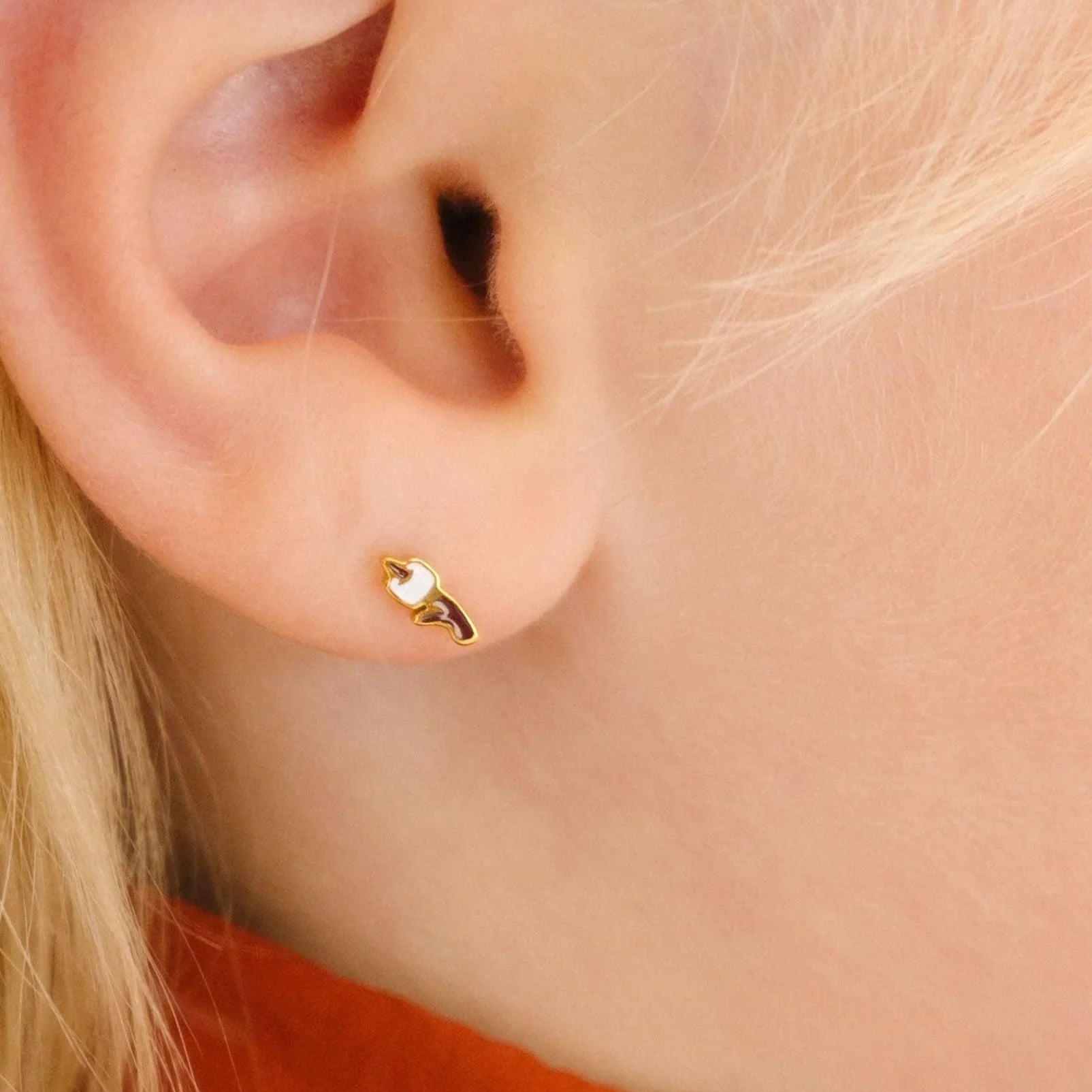 Close up of a blond person's ear with a gold-colored stud earring with a brown and white enamel design that looks like a marshmallow on a stick.