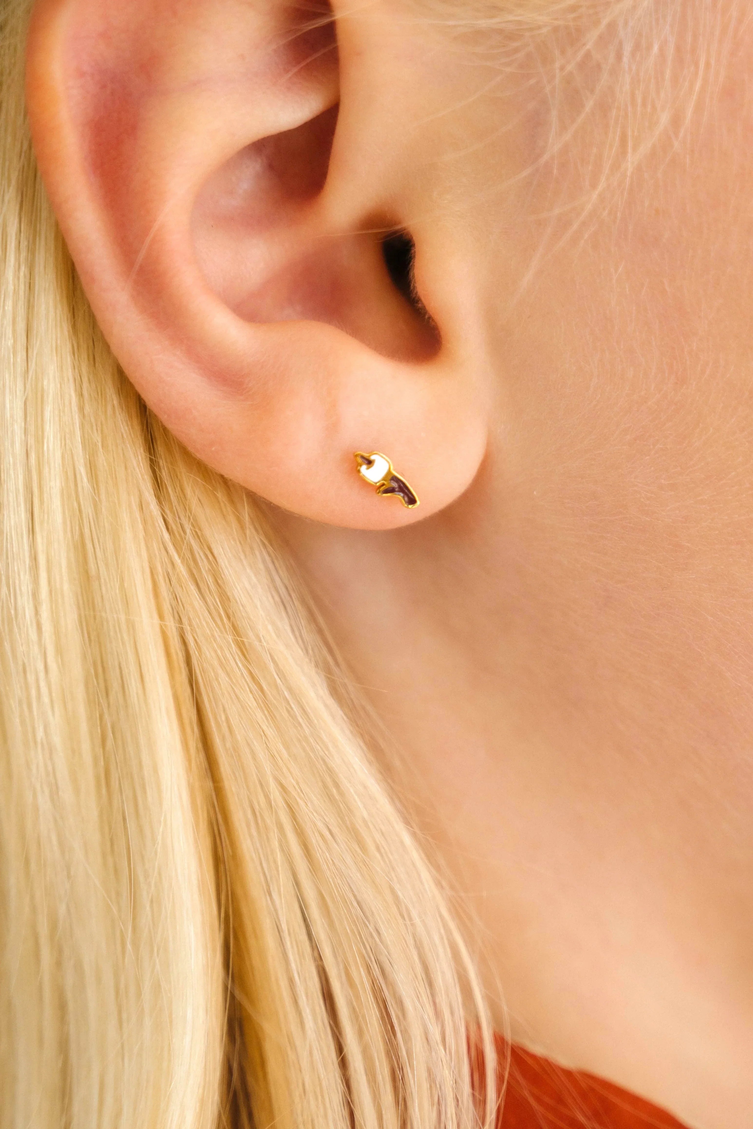 Close up of an ear with a gold-colored stud earring with a brown and white enamel design that looks like a marshmallow on a stick. Blond hair and red shirt collar.
