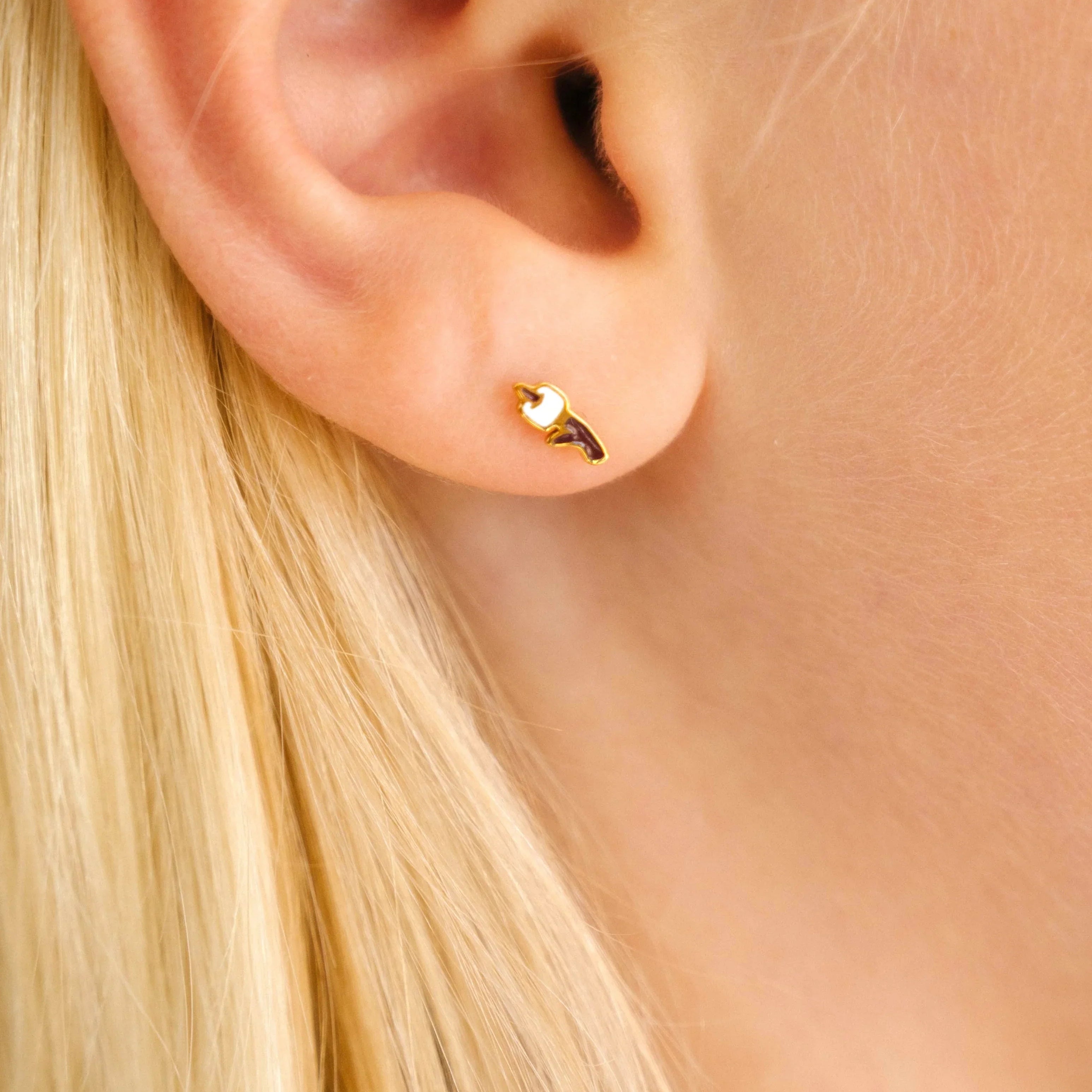 Close up of an ear with a gold-colored stud earring with a brown and white enamel design that looks like a marshmallow on a stick. Blond hair and red shirt collar.