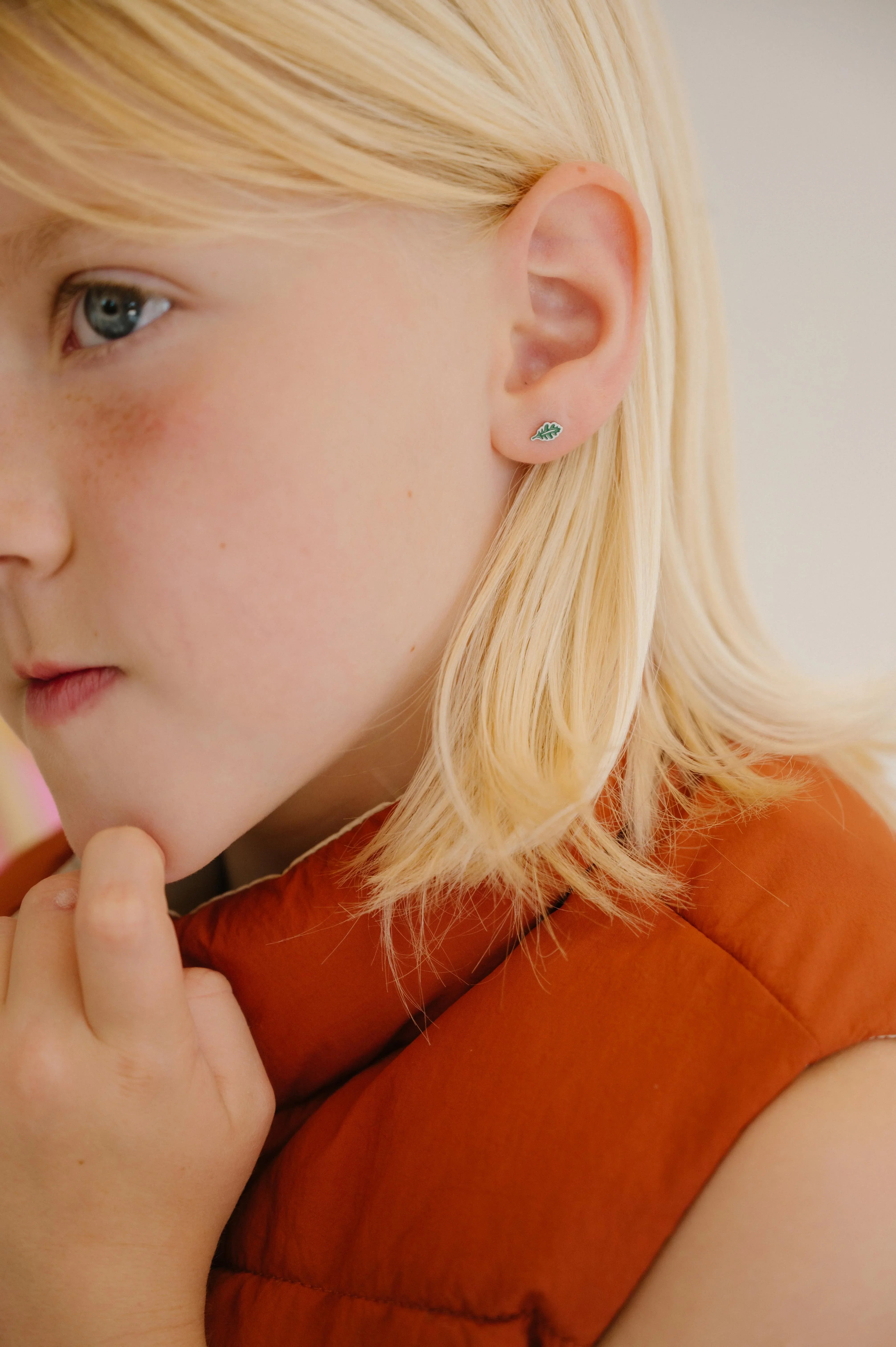 Close-up of a young girl with blonde hair wearing a red vest, and a green oak leaf-shaped enamel stud earring.