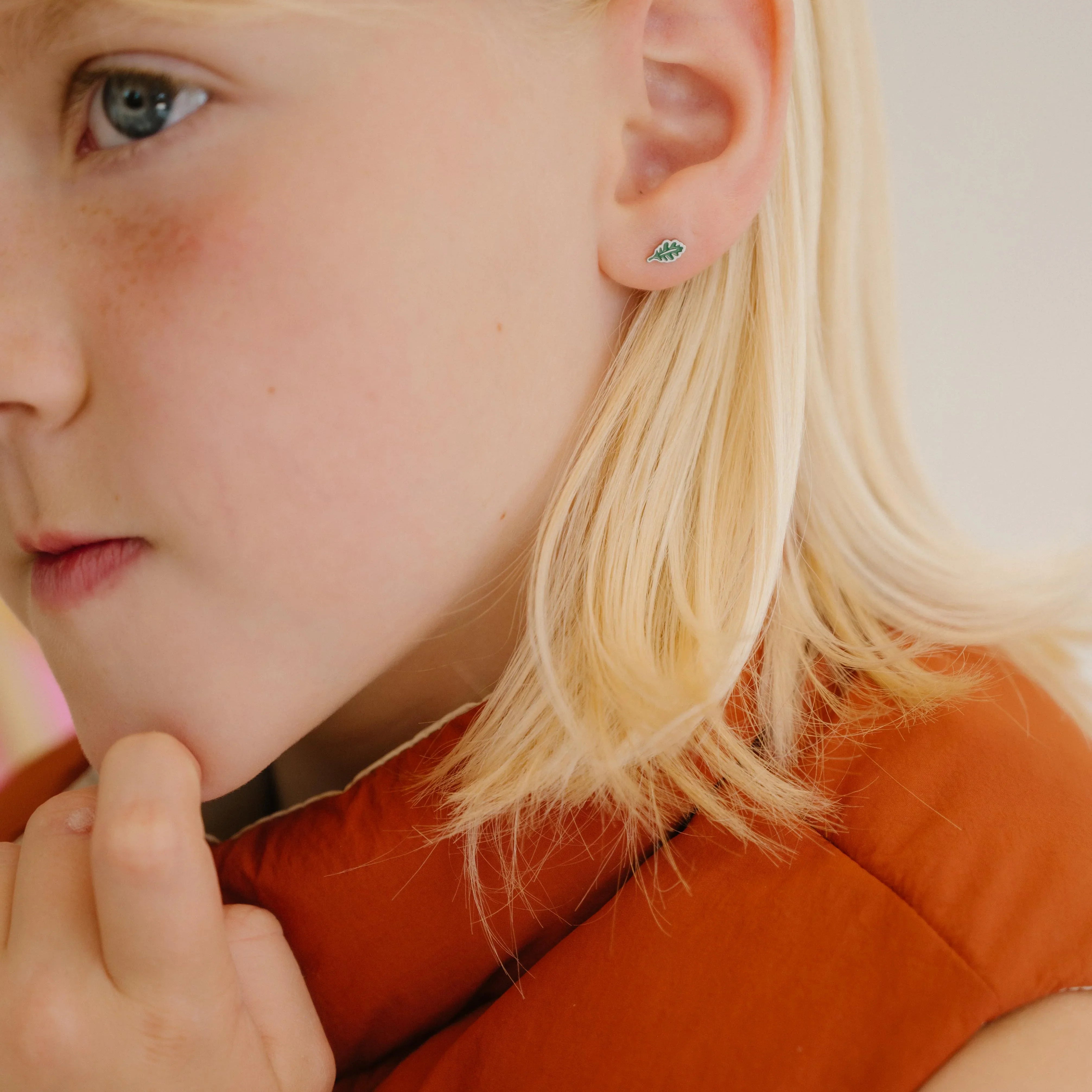 Close-up of a young girl with blonde hair wearing a red vest, and a green oak leaf-shaped enamel stud earring.