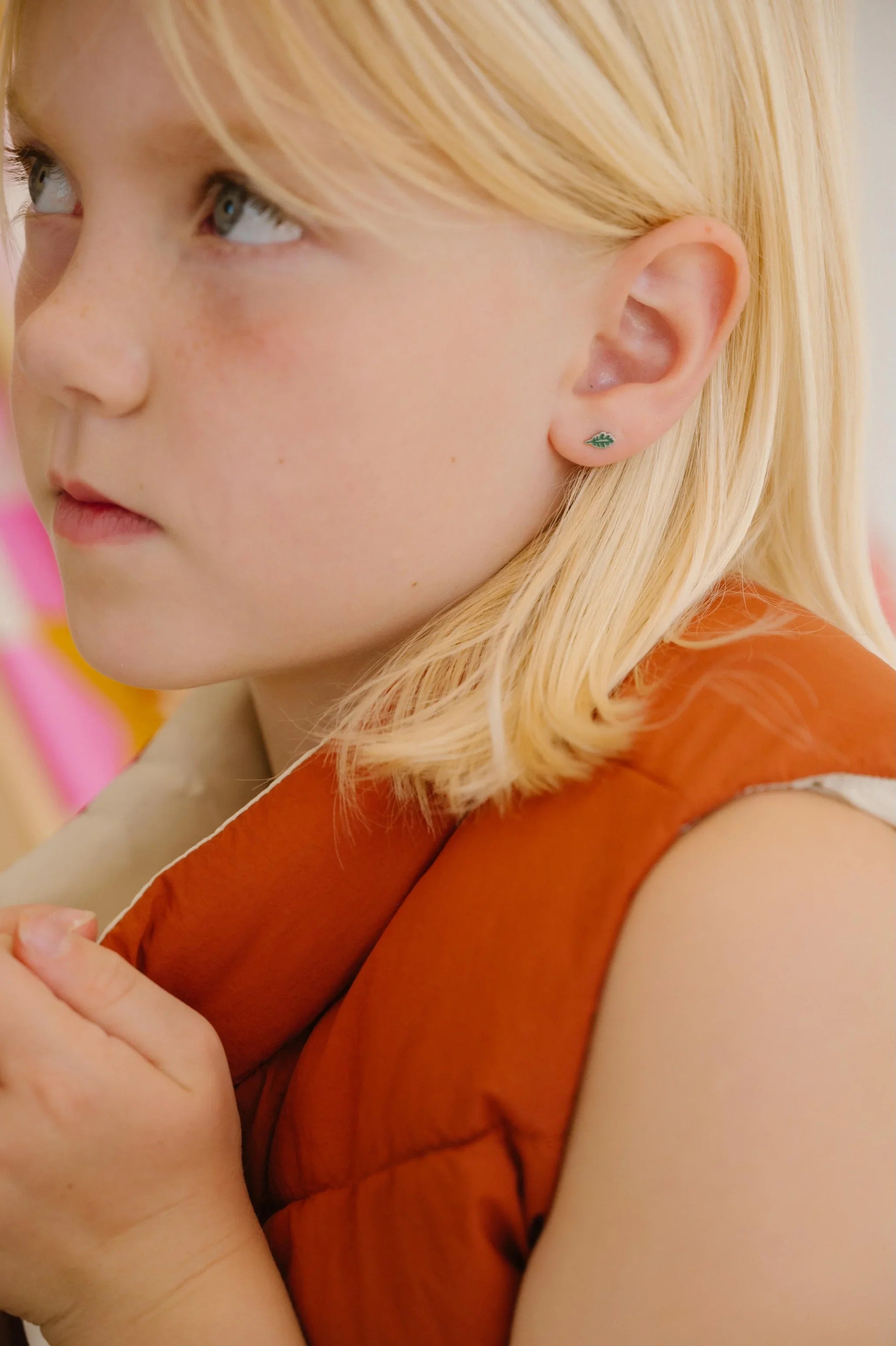 Close-up of a young girl with blonde hair wearing a red vest, and a green oak leaf-shaped enamel stud earring.