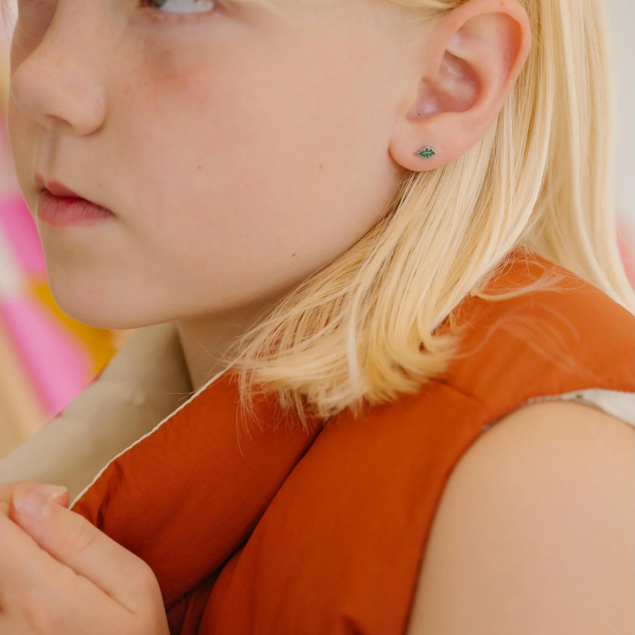 Close-up of a young girl with blonde hair wearing a red vest, and a green oak leaf-shaped enamel stud earring.