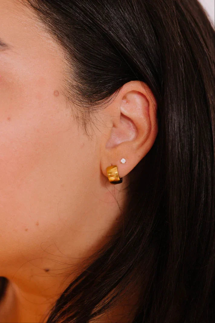 Close-up of an ear with a chunky gold hoop earring and a small pronged opal stud earring.