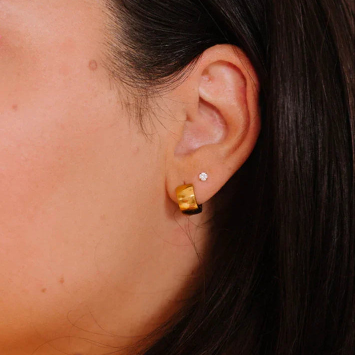 Close-up of an ear with a chunky gold hoop earring and a small pronged opal stud earring.