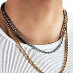 A close-up of a man's neck and chest wearing three layered chain necklaces: a gold curb chain and a silver curb chain, styled with a white ribbed tank top.
