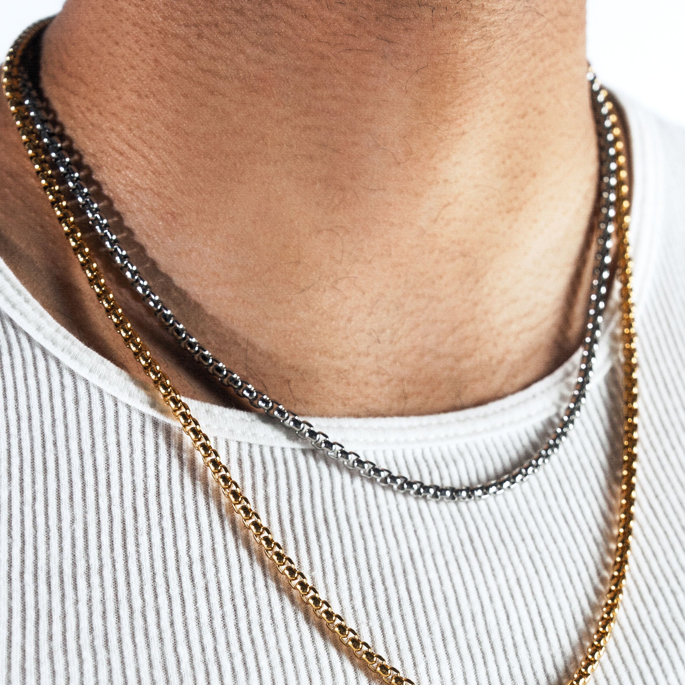 A close-up of a man's neck and chest wearing three layered chain necklaces: a gold curb chain and a silver curb chain, styled with a white ribbed tank top.