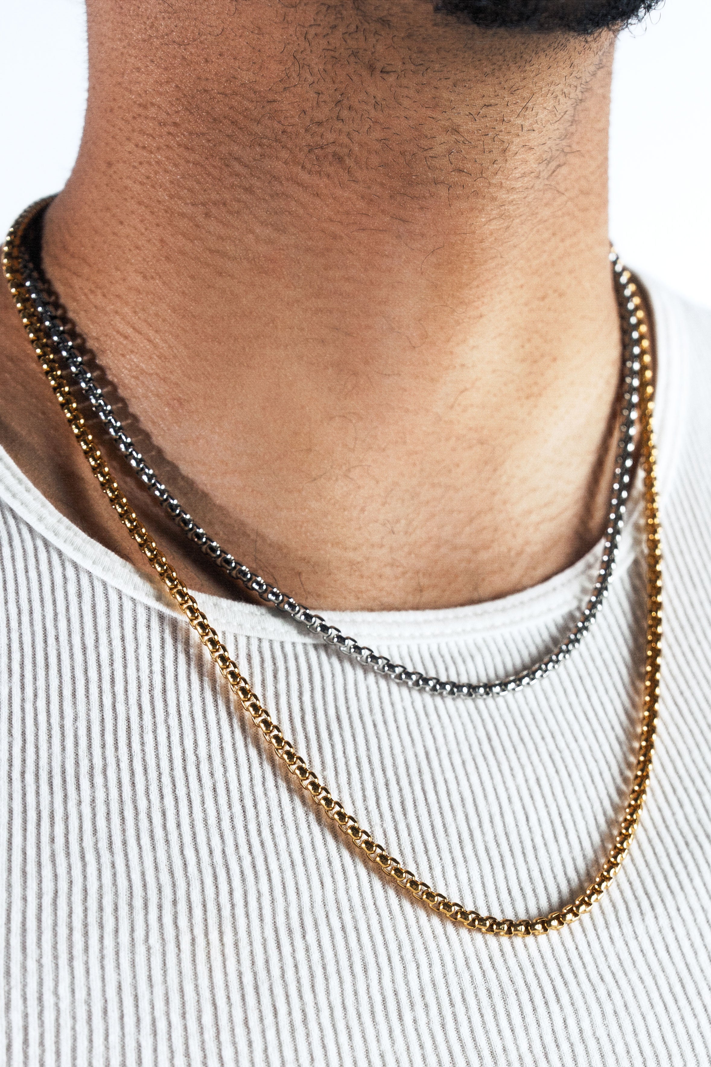 A close-up of a man's neck and chest wearing three layered chain necklaces: a gold curb chain and a silver curb chain, styled with a white ribbed tank top.