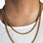A close-up of a person's neck and upper chest wearing a double-layered gold chain necklace over a white ribbed tank top.