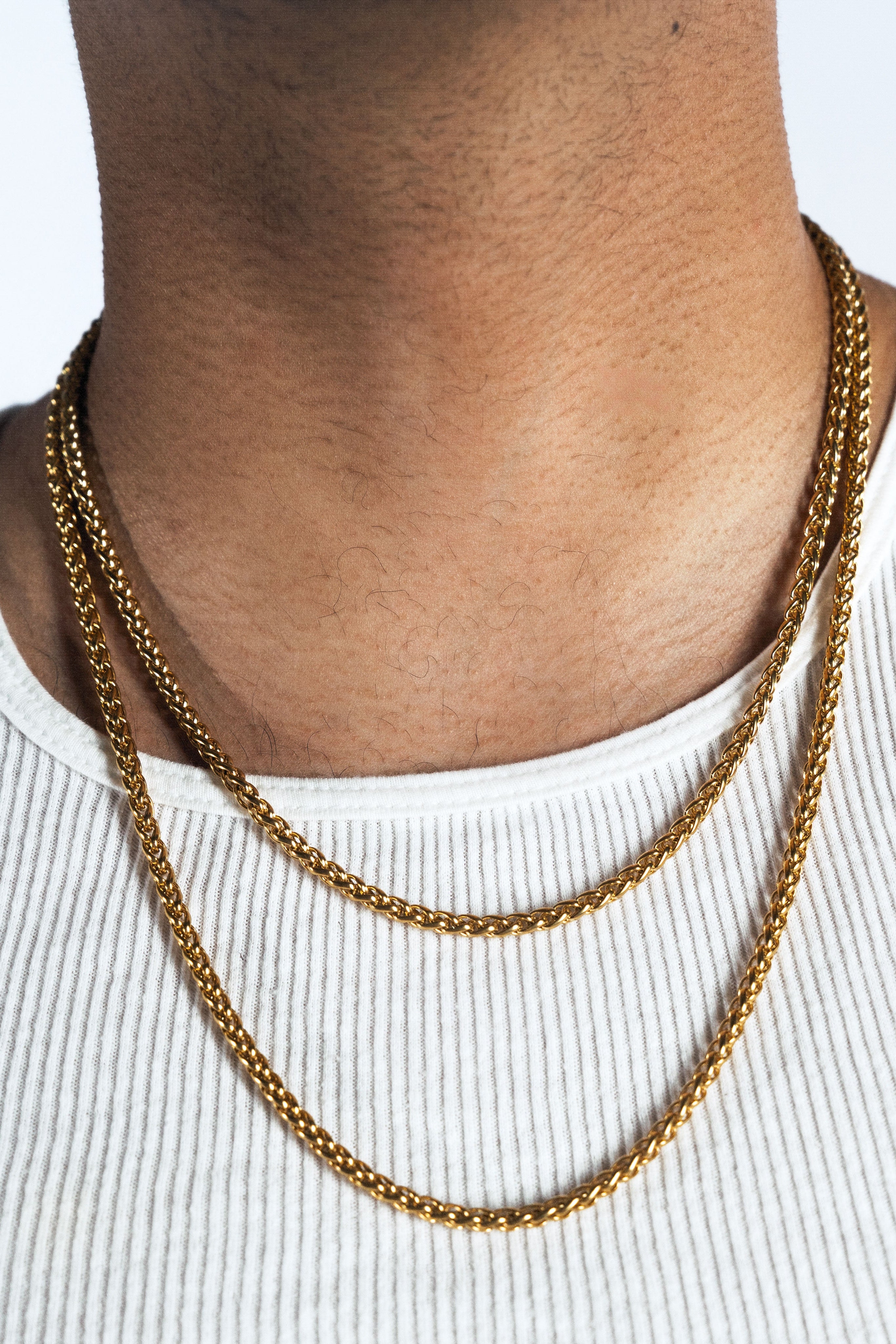 A close-up of a person's neck and upper chest wearing a double-layered gold chain necklace over a white ribbed tank top.