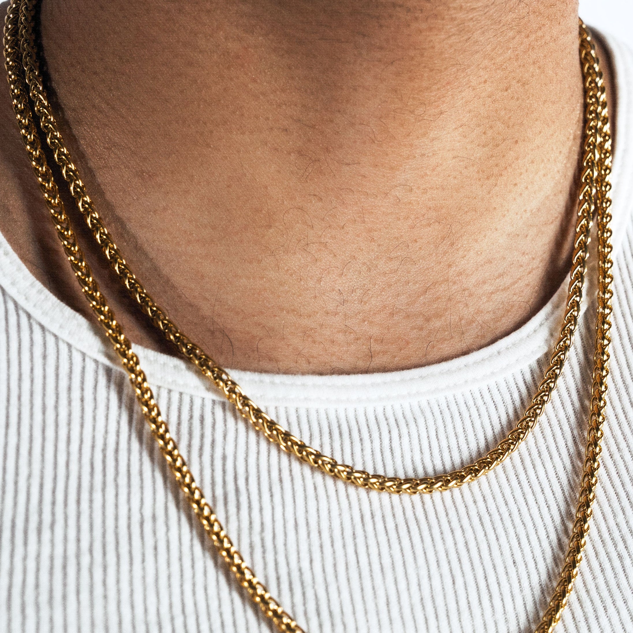 A close-up of a man wearing a layered gold chain necklace over a white ribbed tank top, with a white background.