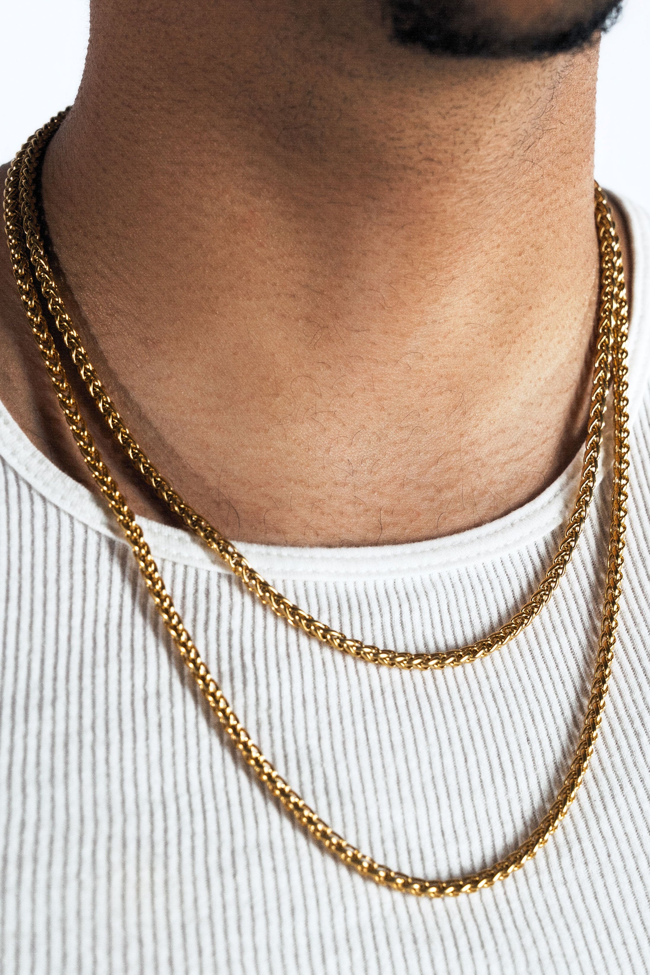 A close-up of a man wearing a layered gold chain necklace over a white ribbed tank top, with a white background.