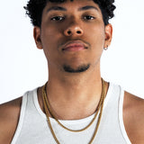 A man with curly black hair wearing a white ribbed tank top and two gold chain necklaces, photographed against a plain white studio background.