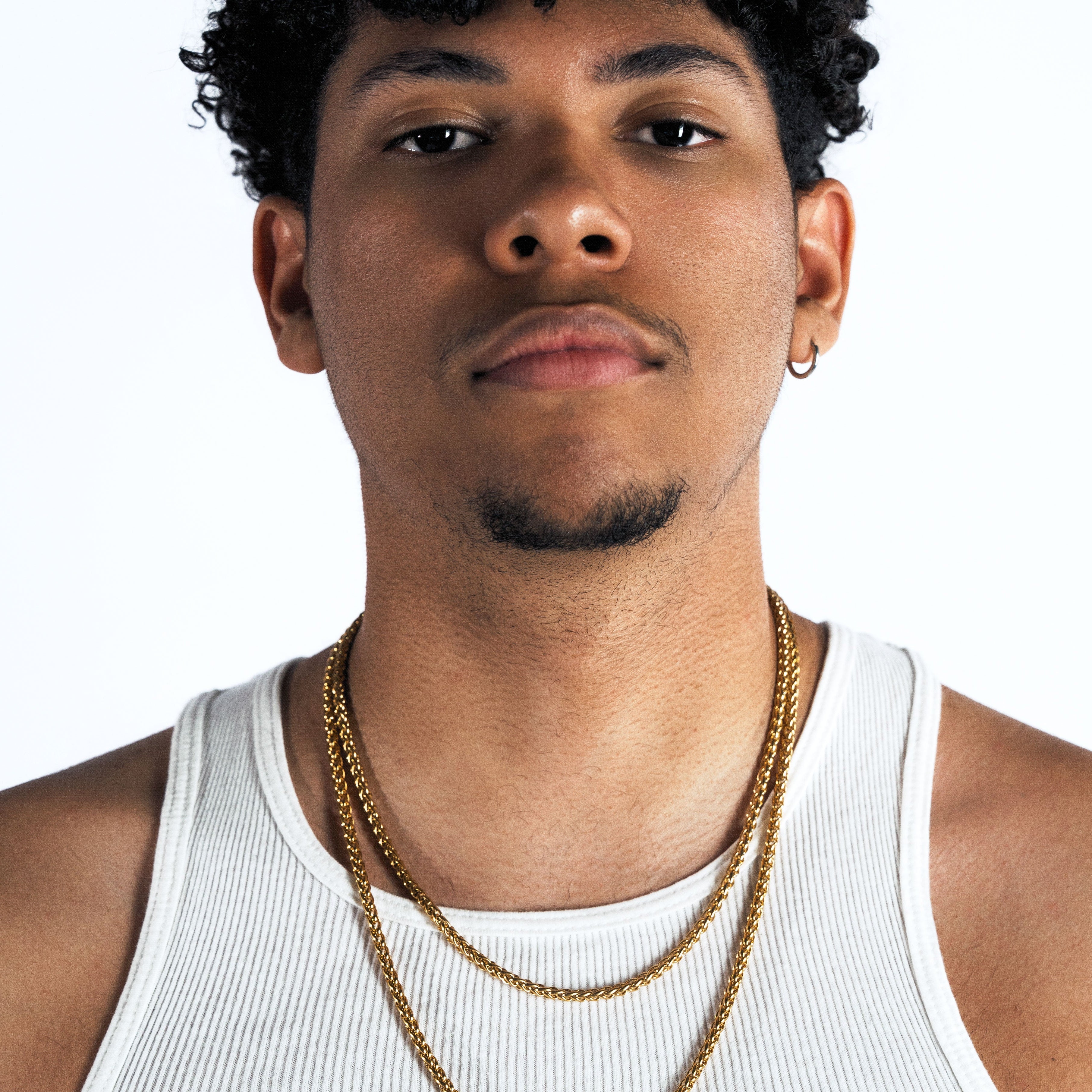 A man with curly black hair wearing a white ribbed tank top and two gold chain necklaces, photographed against a plain white studio background.