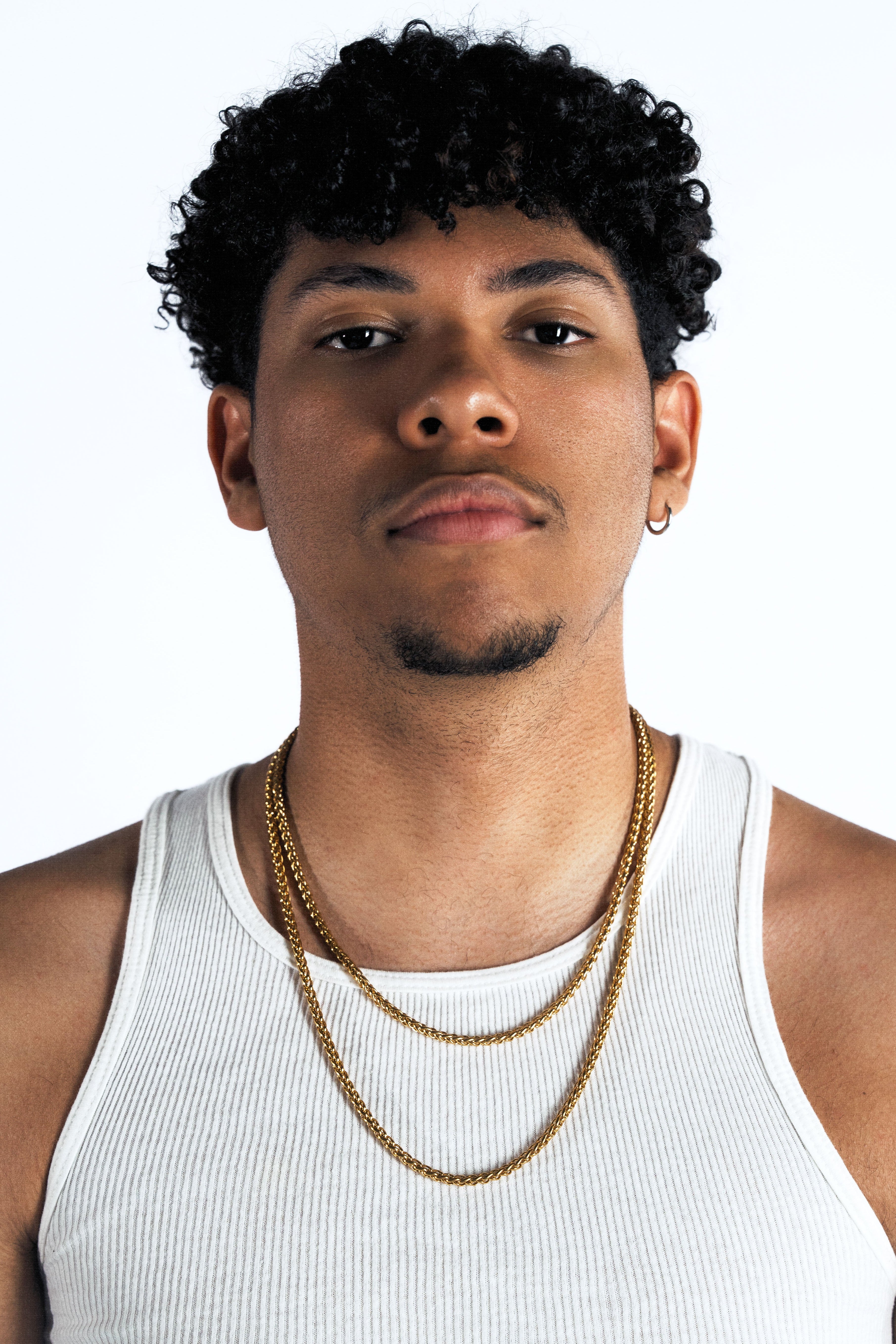 A man with curly black hair wearing a white ribbed tank top and two gold chain necklaces, photographed against a plain white studio background.