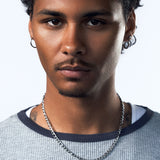 A close-up portrait of a man with curly black hair, wearing a silver chain necklace and hoop earrings, wearing a gray textured ringer t-shirt with a dark navy collar, against a light gray background.