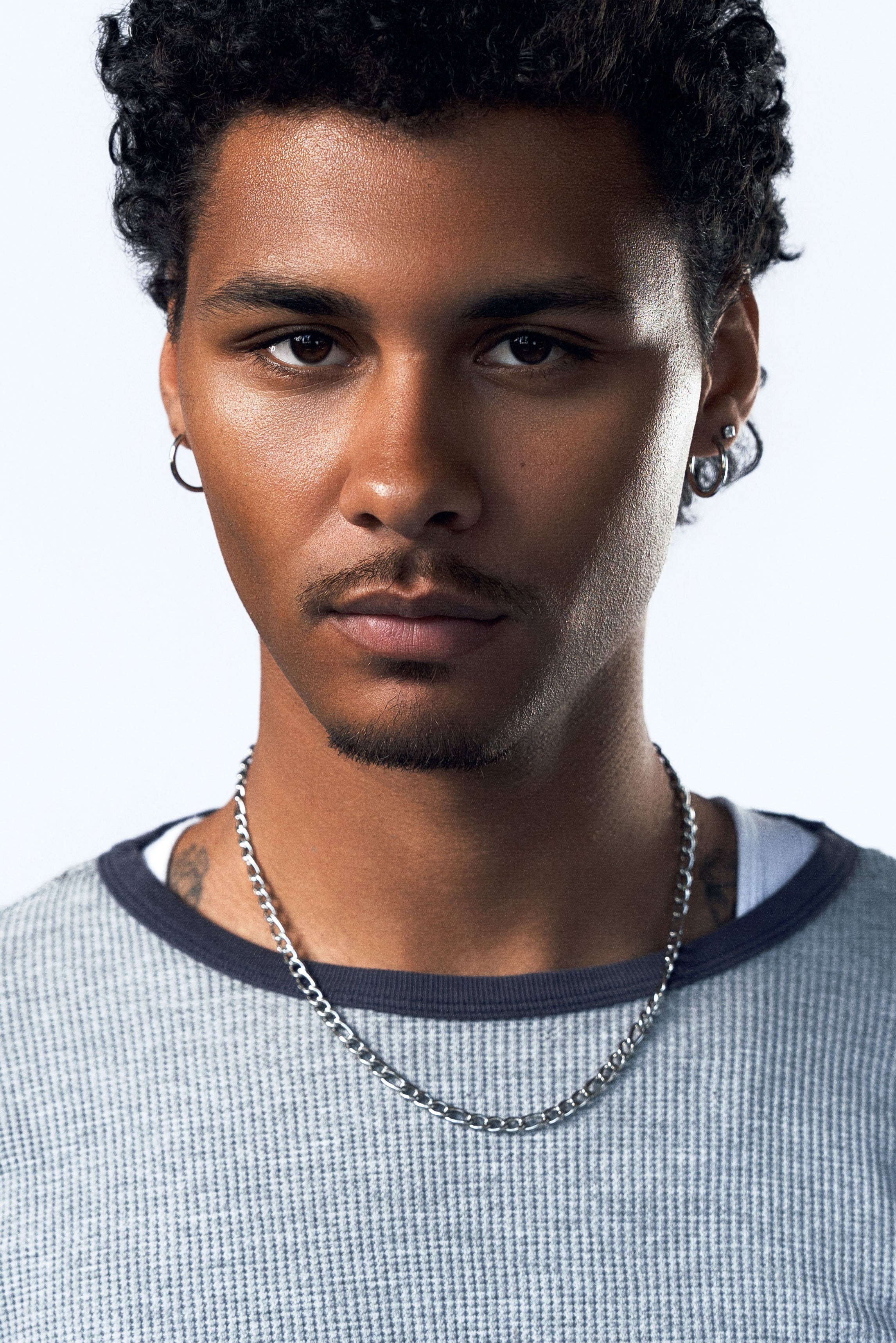 A close-up portrait of a man with curly black hair, wearing a silver chain necklace and hoop earrings, wearing a gray textured ringer t-shirt with a dark navy collar, against a light gray background.
