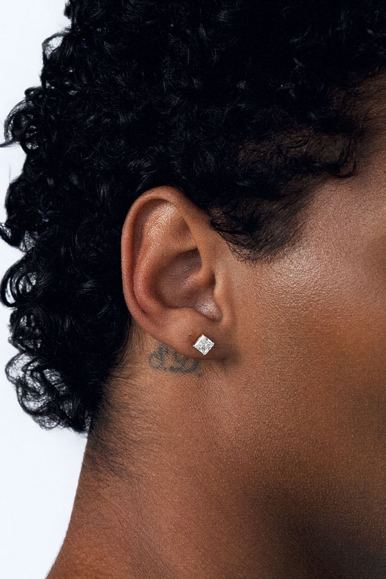 A close-up side profile of a person with curly black hair wearing a small, clear square-cut CZ stud earring on their earlobe, against a plain white background.