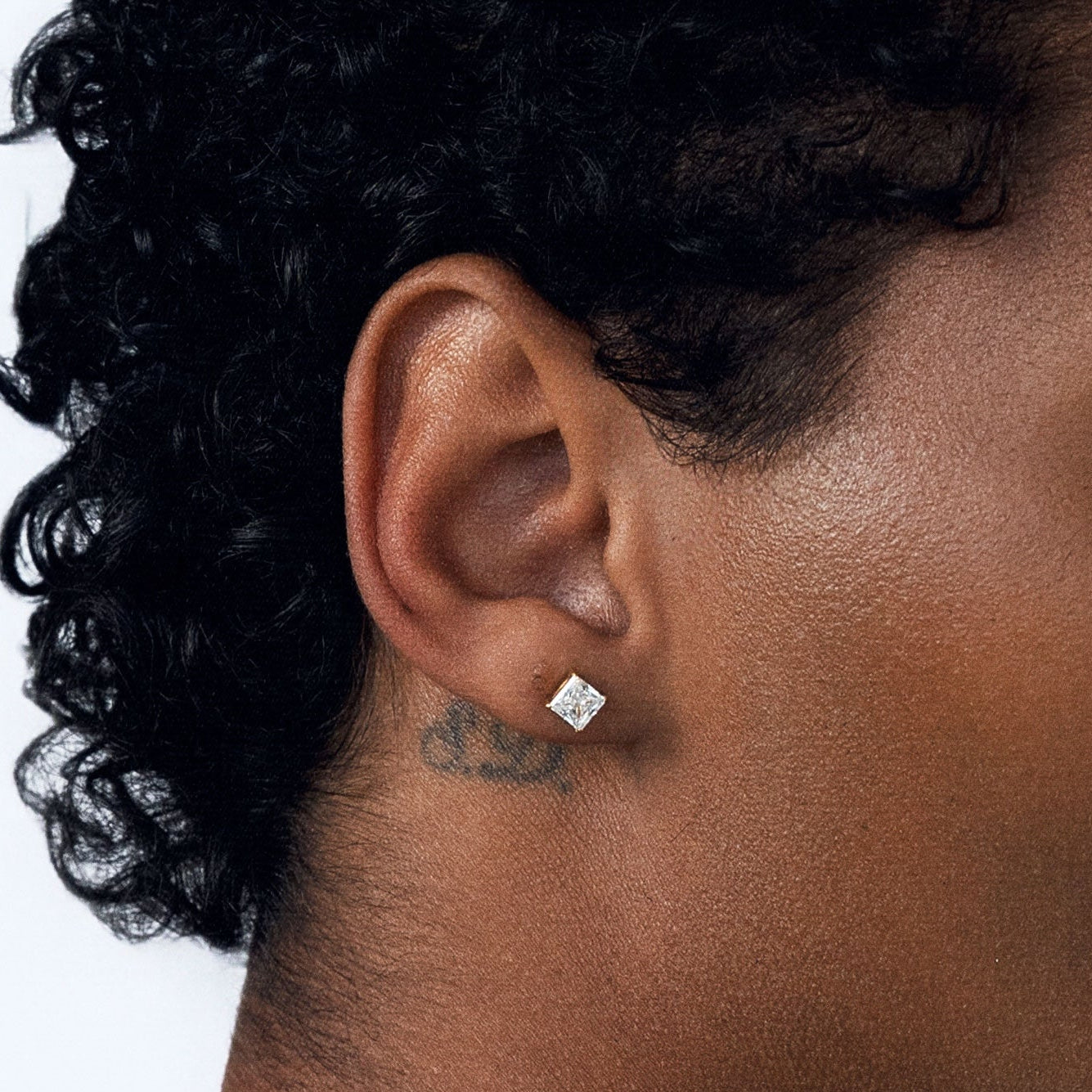 A close-up side profile of a person with curly black hair wearing a small, clear square-cut CZ stud earring on their earlobe, against a plain white background.
