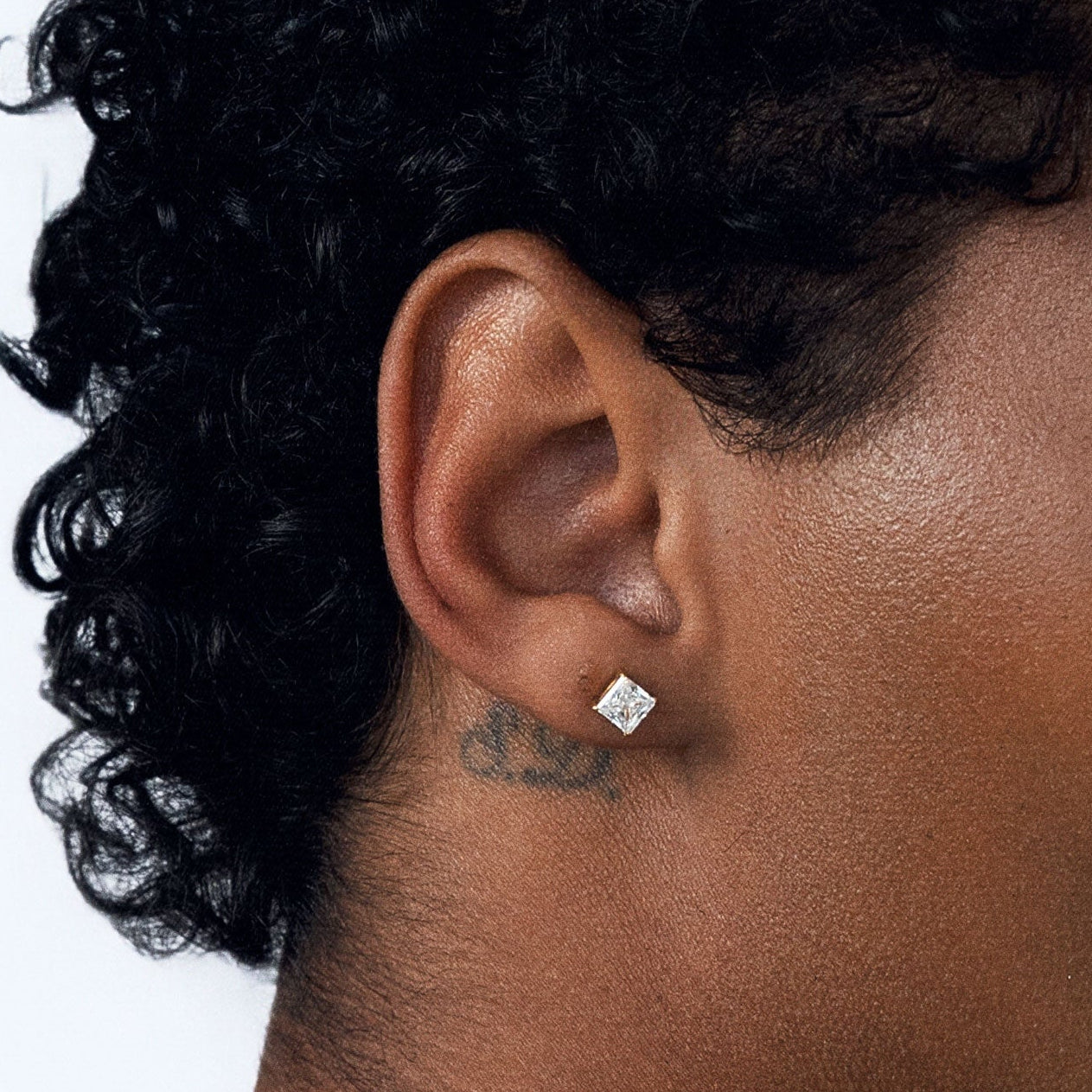A close-up side profile of a person with curly black hair wearing a statement-sized, clear square-cut CZ stud earring on their earlobe, against a plain white background.