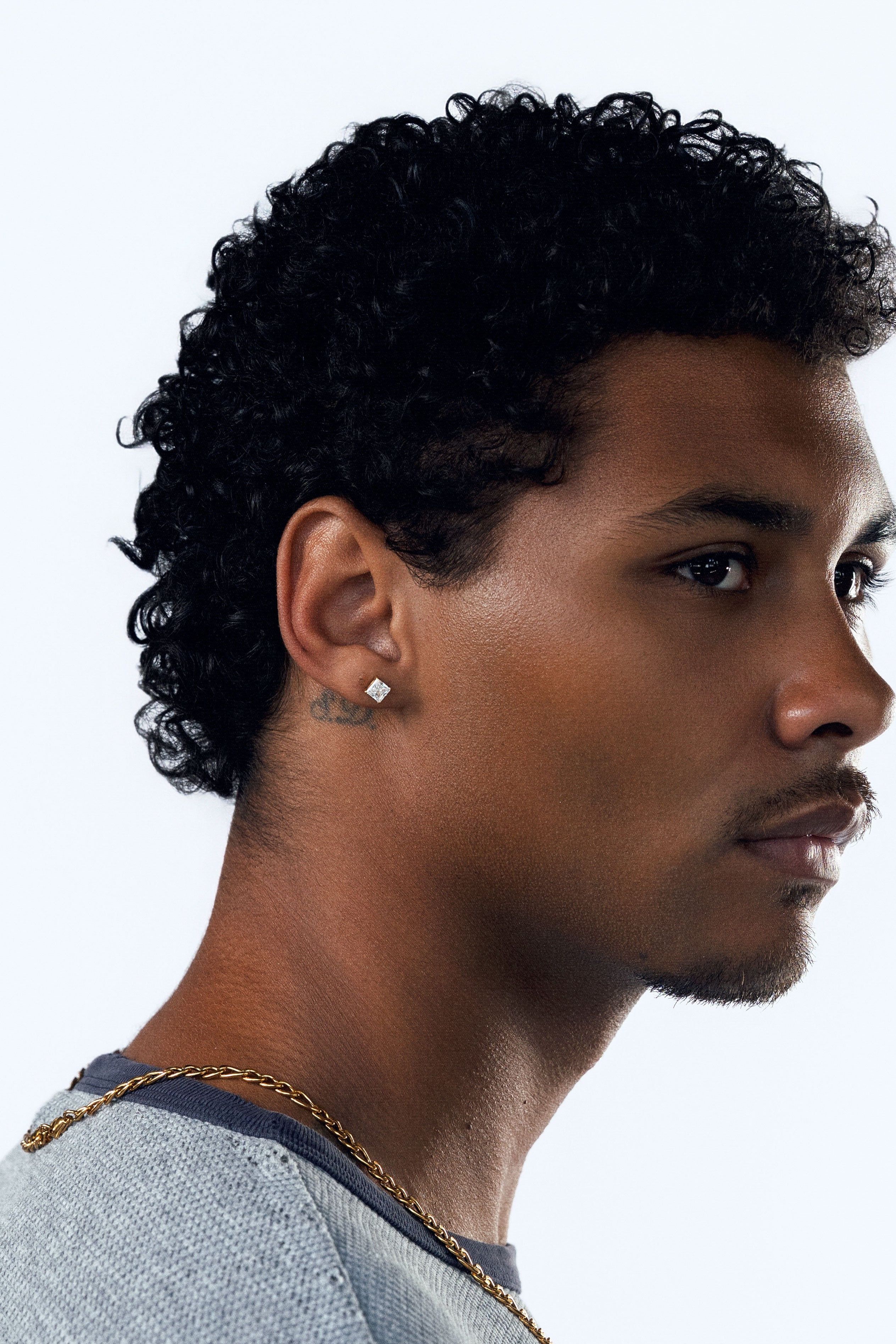 A man in profile wearing a light blue-gray textured crewneck shirt, a gold chain necklace, and a square CZ stud earring, set against a plain white studio background.