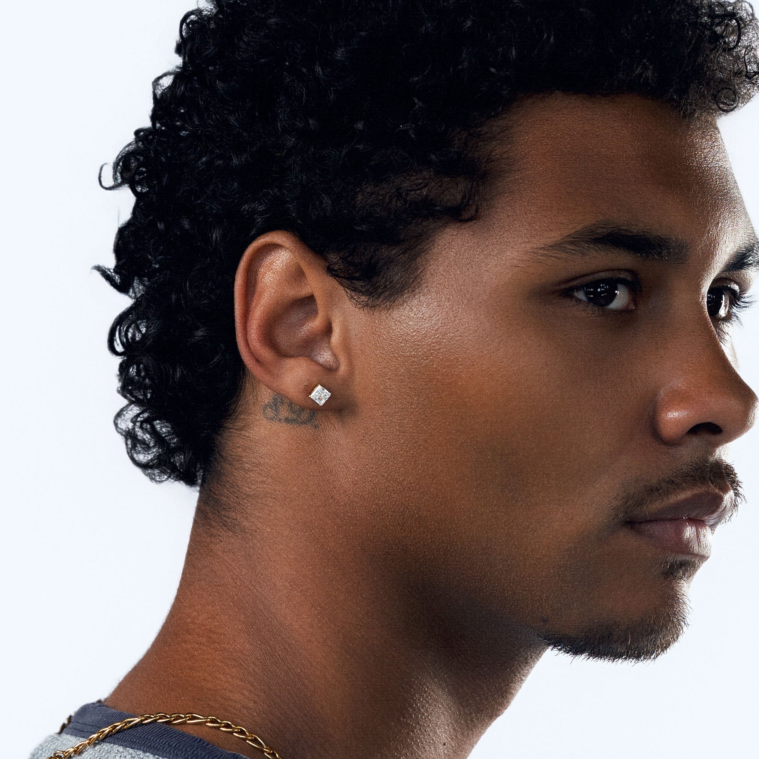 A man in profile wearing a light blue-gray textured crewneck shirt, a gold chain necklace, and a square CZ stud earring, set against a plain white studio background.