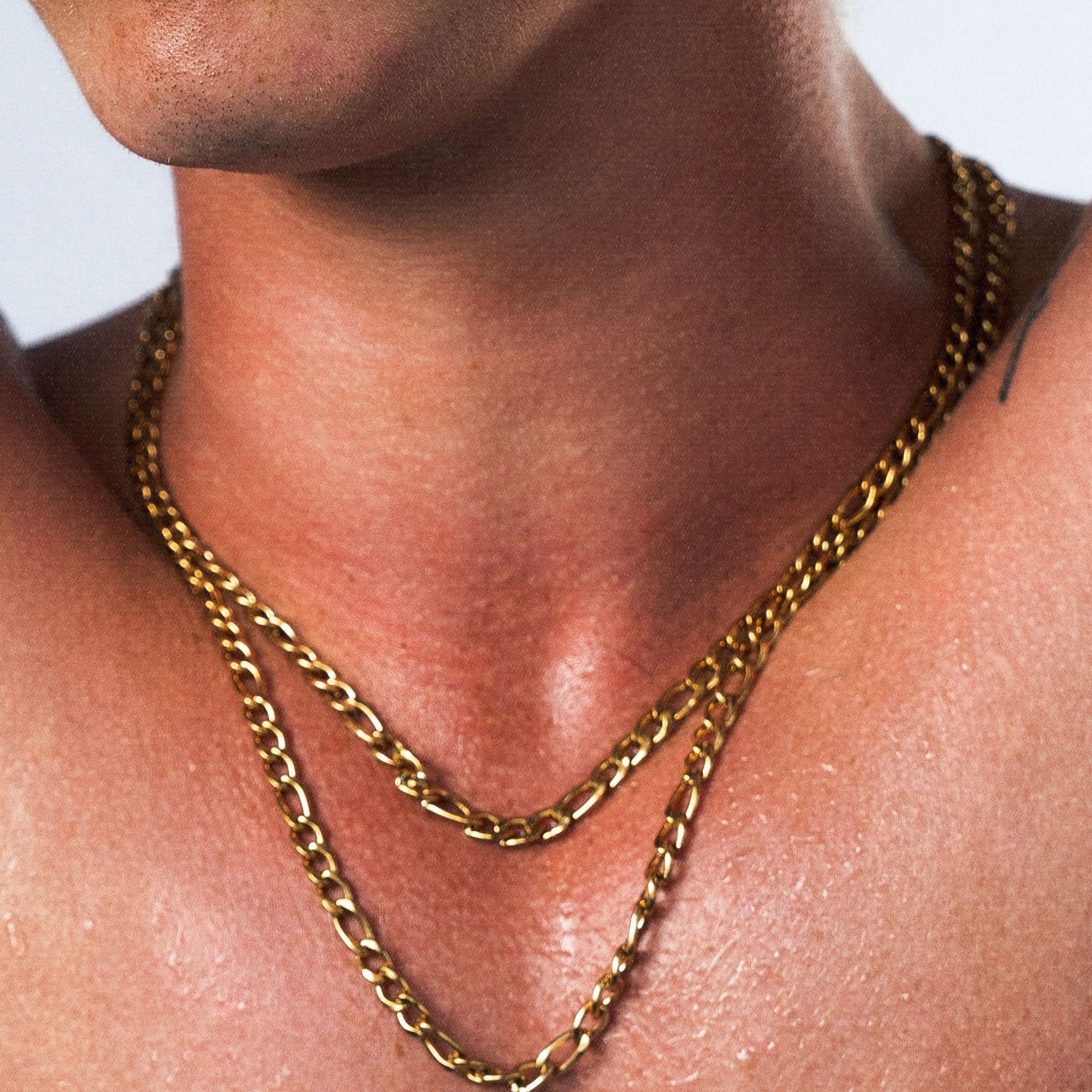 A shirtless man with dark hair poses with his arms behind his head, wearing two gold chain necklaces against a light gray studio background.
