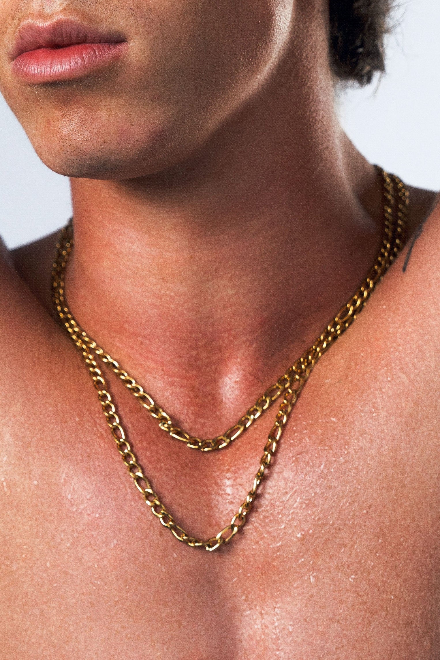A shirtless man with dark hair poses with his arms behind his head, wearing two gold chain necklaces against a light gray studio background.