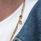 A close-up of a person wearing a delicate gold chain necklace with a small rectangular charm, styled over a white t-shirt and blue denim jacket.