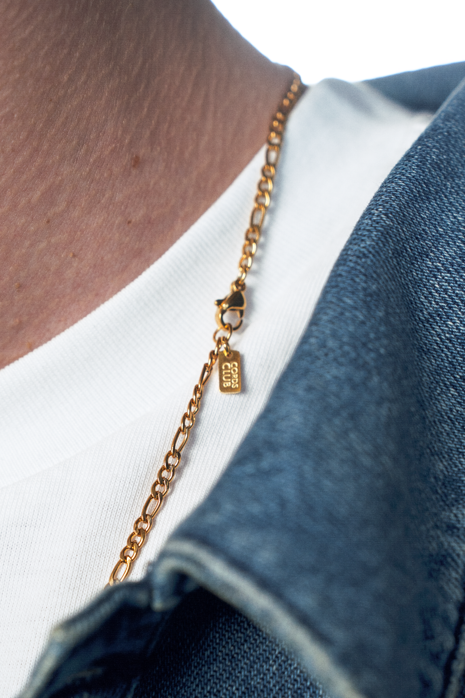 A close-up of a person wearing a delicate gold chain necklace with a small rectangular charm, styled over a white t-shirt and blue denim jacket.