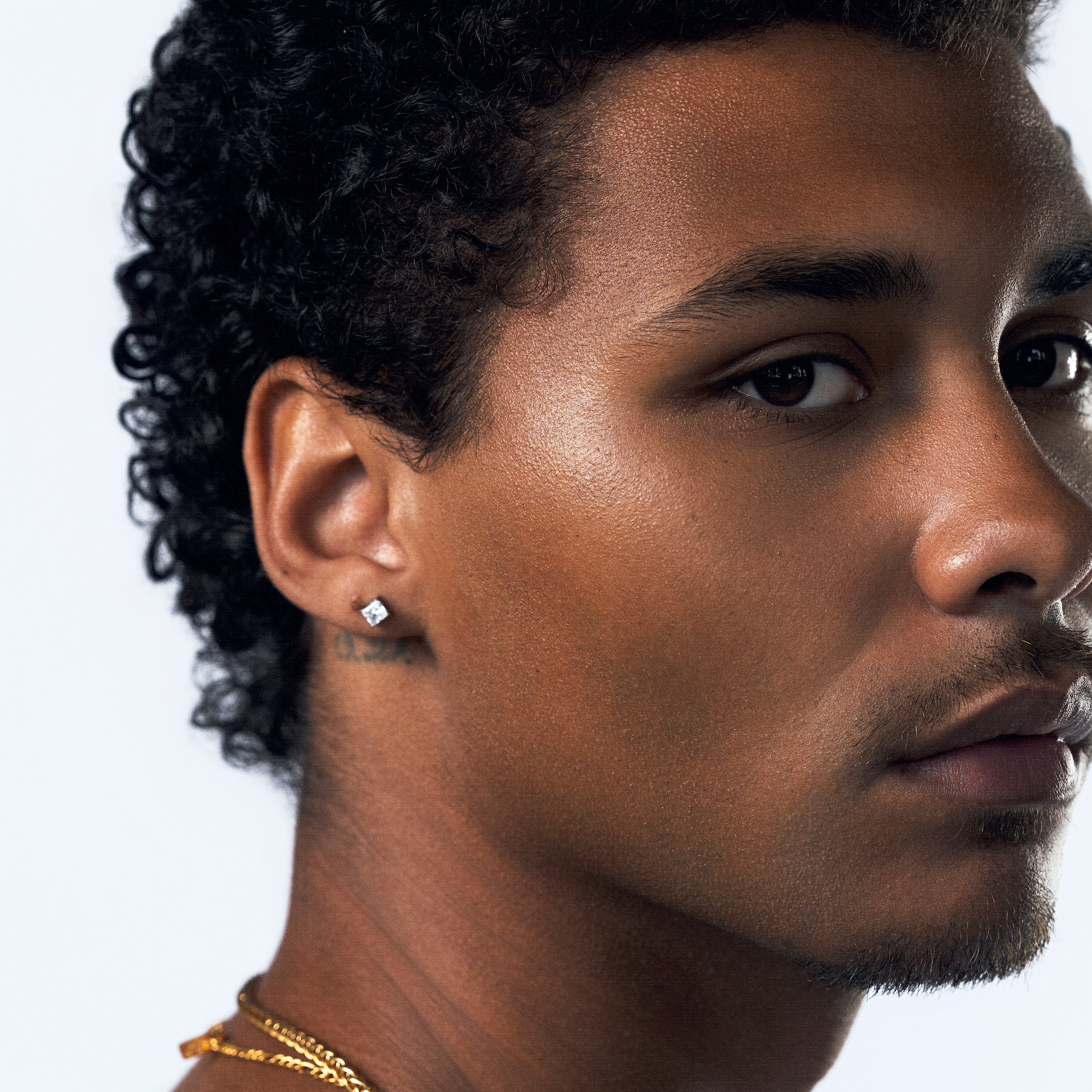 A close-up portrait of a man with curly black hair, wearing a white tank top, a gold chain necklace, and a small square cz stud earring, shown against a plain light gray background.