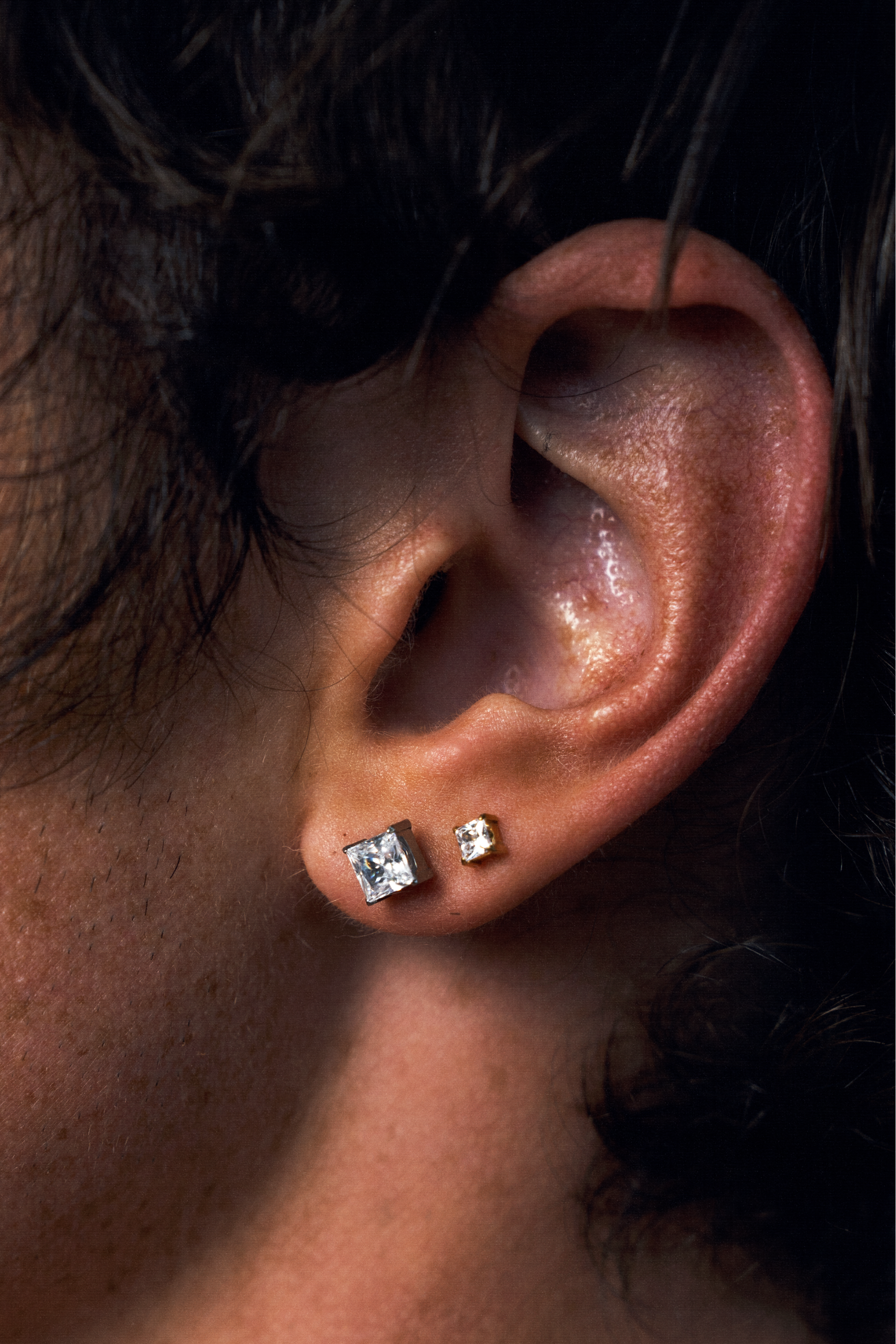 Close-up of a person's ear wearing two small square stud earrings, one larger and one smaller, both with clear CZ gemstones in a gold-tone setting.