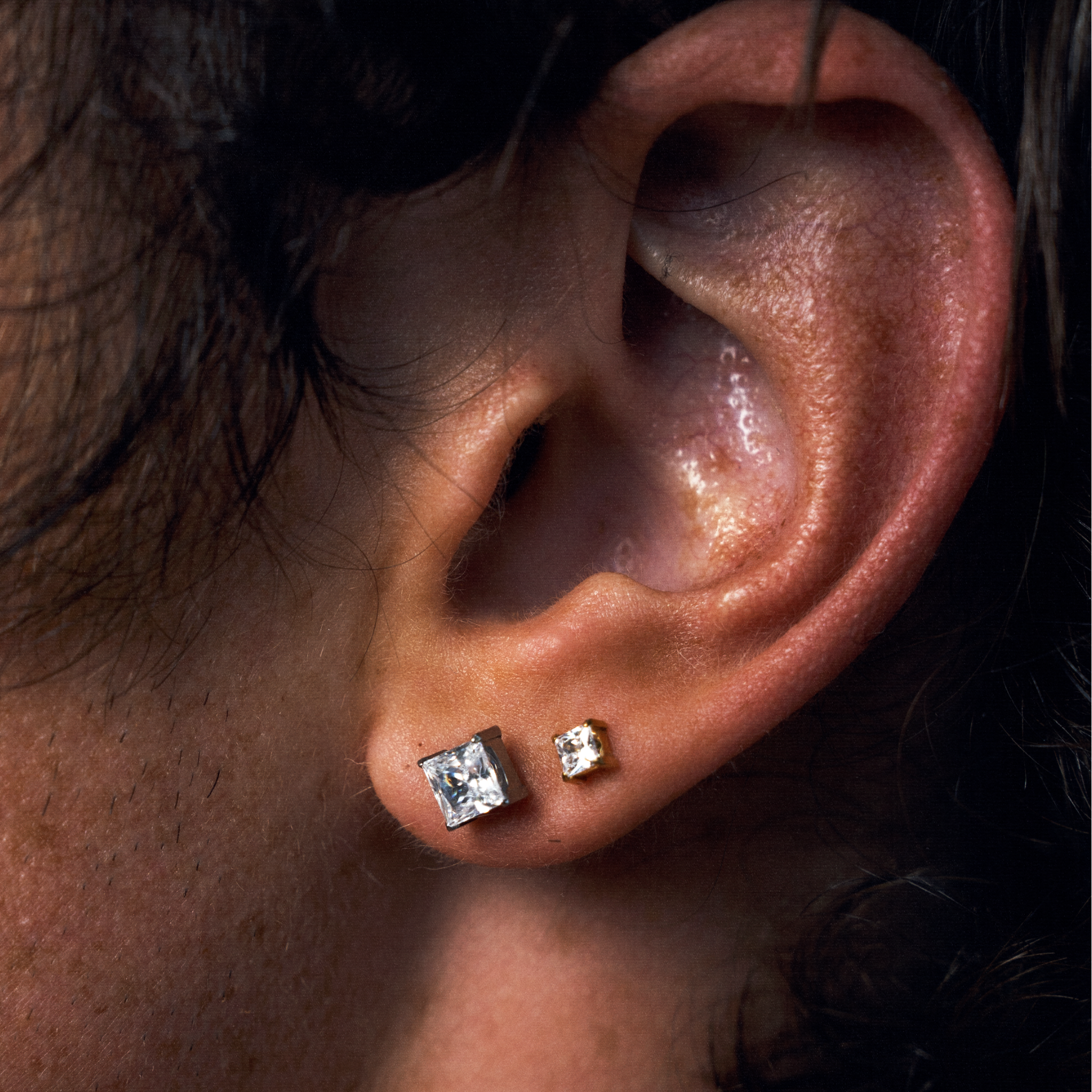 Close-up of a person's ear wearing two small square stud earrings, one larger and one smaller, both with clear CZ gemstones in a gold-tone setting.