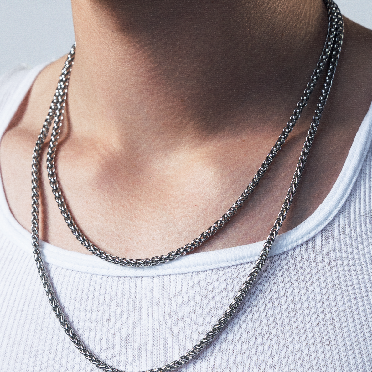 A close-up shot of a young man with curly brown hair wearing a white ribbed tank top and layered silver chain necklaces, set against a plain light gray background.