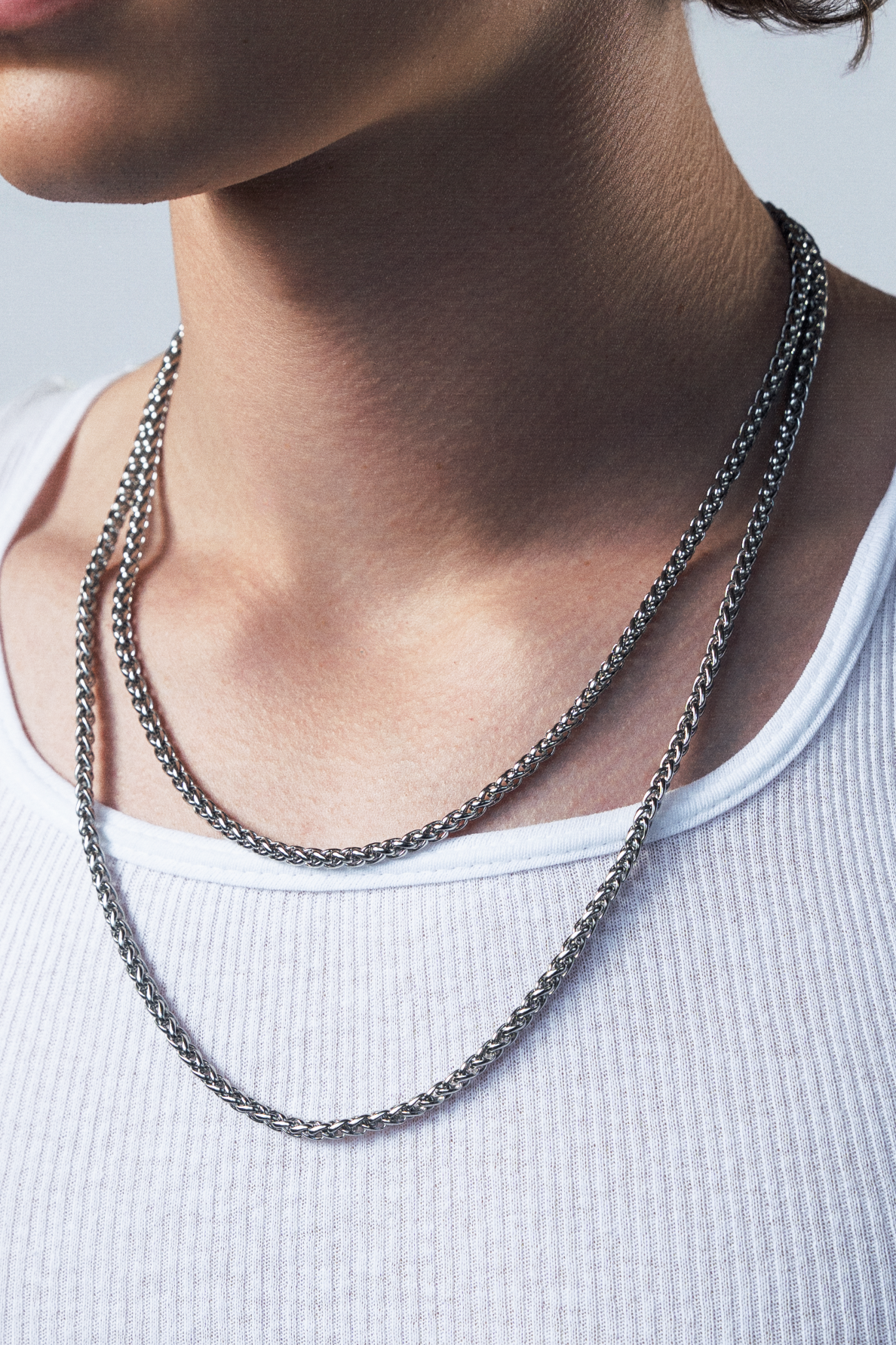 A close-up shot of a young man with curly brown hair wearing a white ribbed tank top and layered silver chain necklaces, set against a plain light gray background.