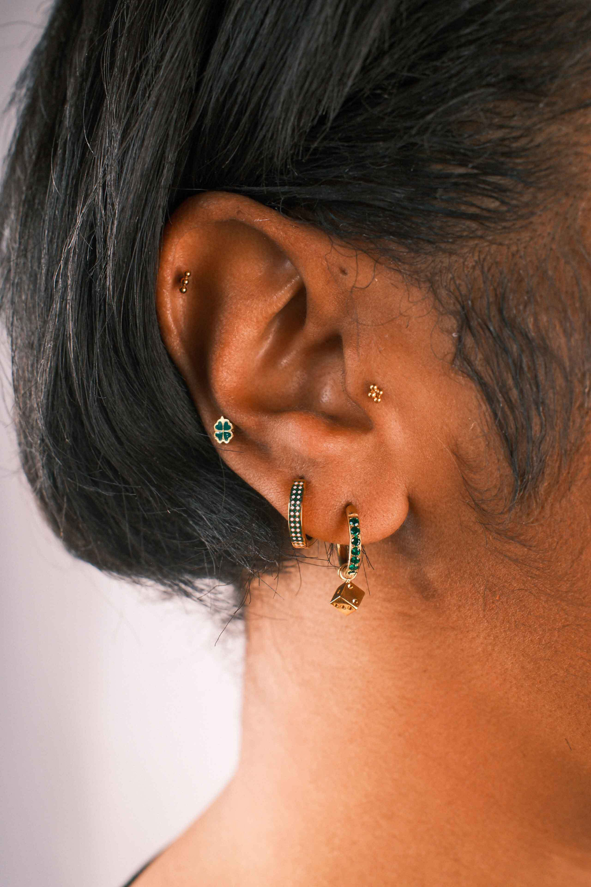 Close-up of an ear wearing gold hoop earrings with green gemstones against a neutral background