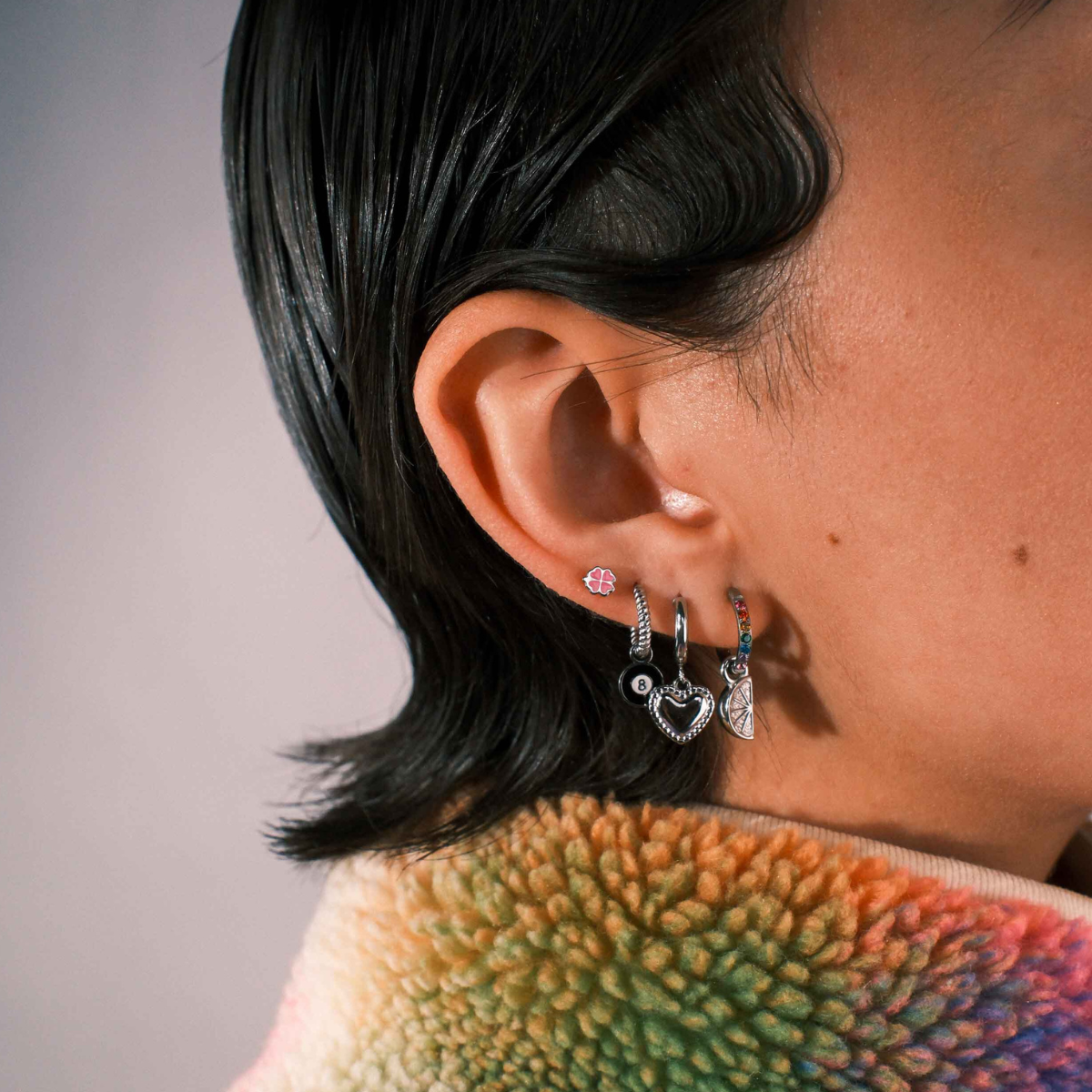 Close-up of a person wearing multiple silver earrings including a clover shaped flat back stud in pink enamel. 