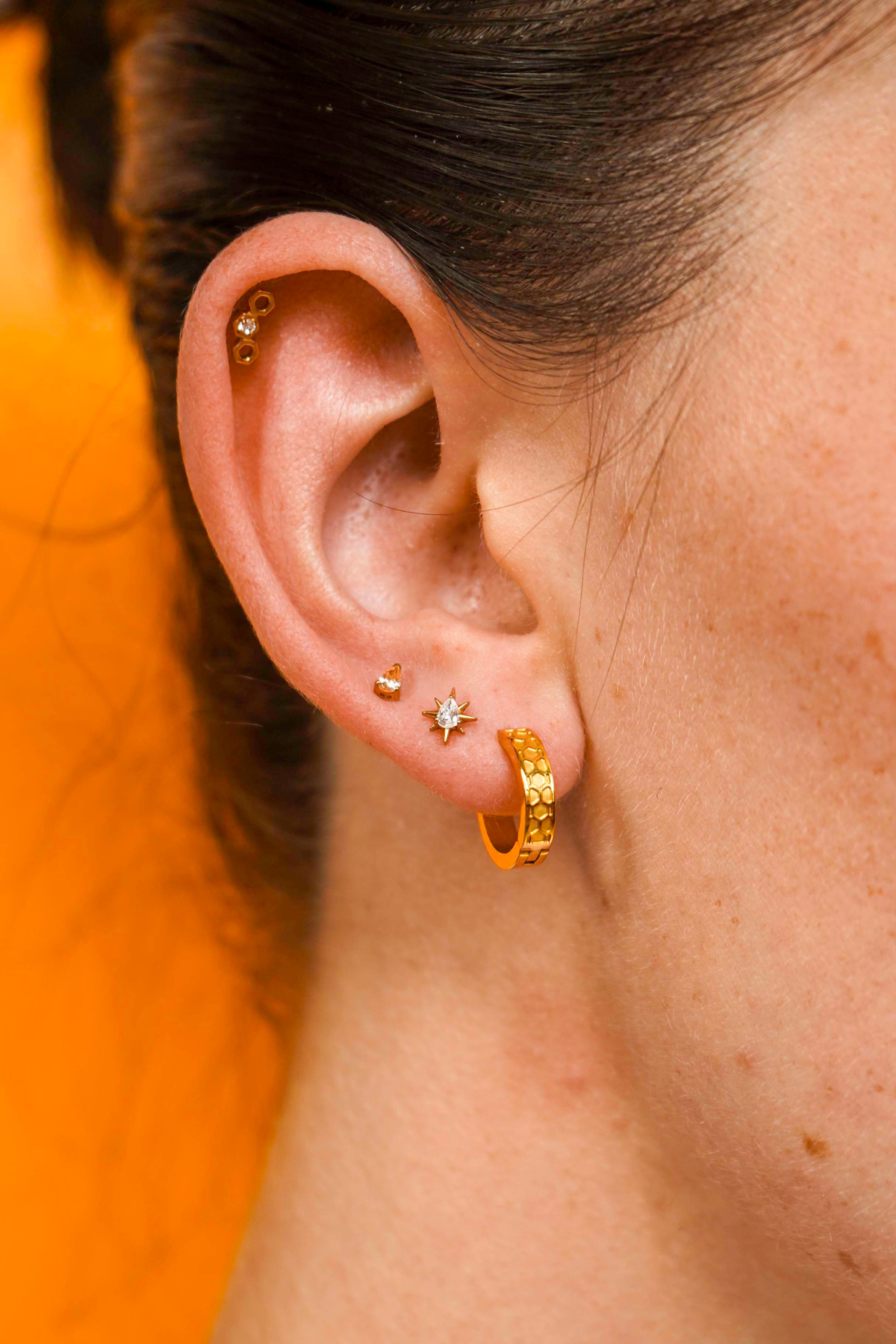 Close up of a model wearing a hexagon shaped flat back stud earring made from 18k gold-plated ASTM 136 implant-grade titanium, with a clear zirconia stone, against a blurred honey-colored background. 