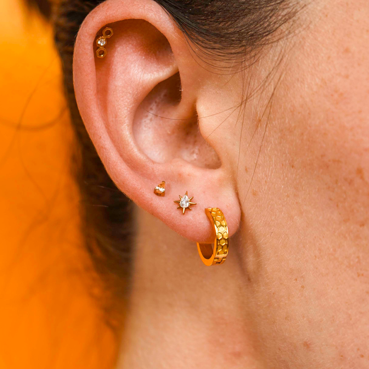 Close up of a model wearing a hexagon shaped flat back stud earring made from 18k gold-plated ASTM 136 implant-grade titanium, with a clear zirconia stone, against a blurred honey-colored background. 