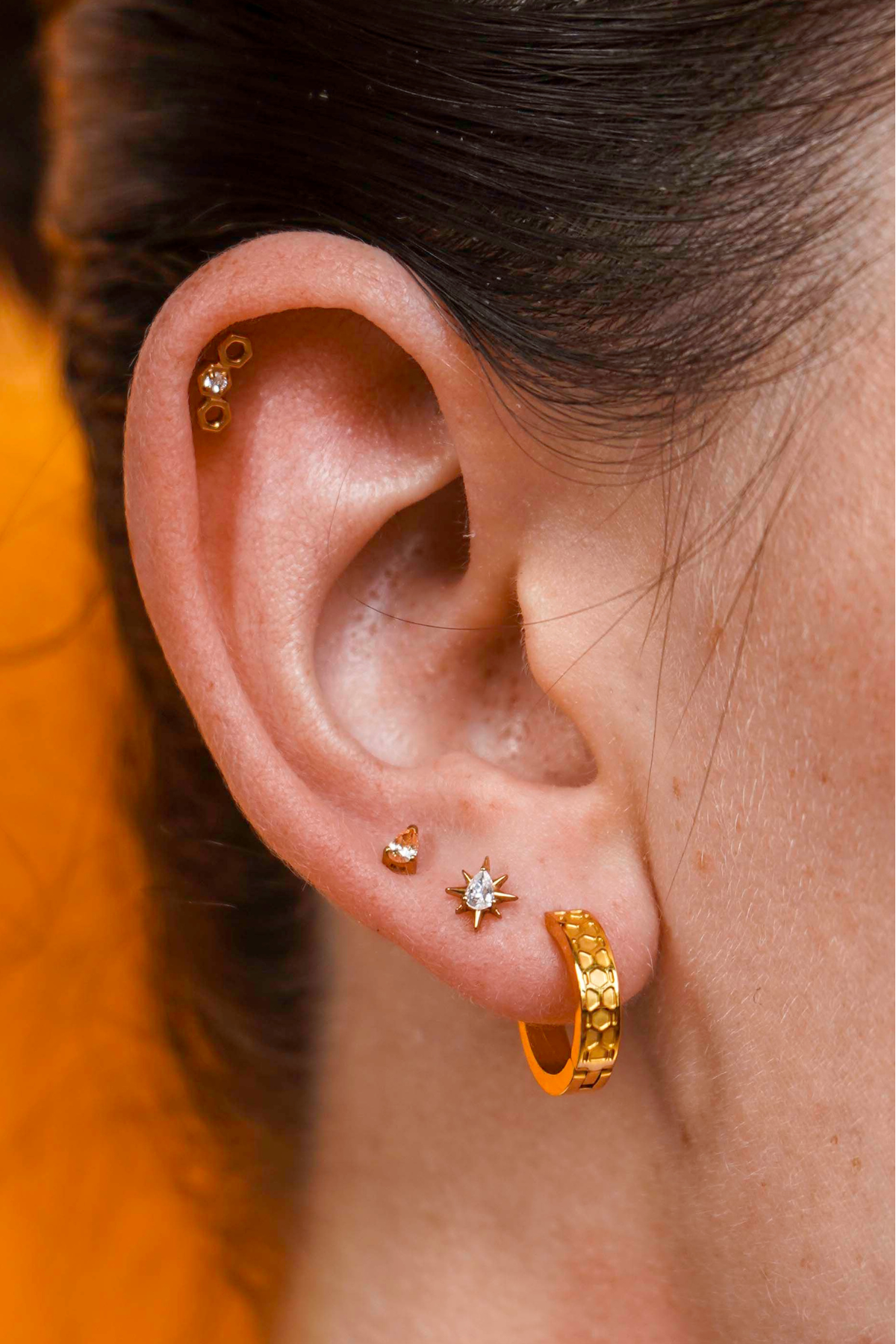 Close up of a model wearing a hexagon shaped flat back stud earring made from 18k gold-plated ASTM F136 implant-grade titanium, with a clear zirconia stone, against a blurred honey-colored background. 