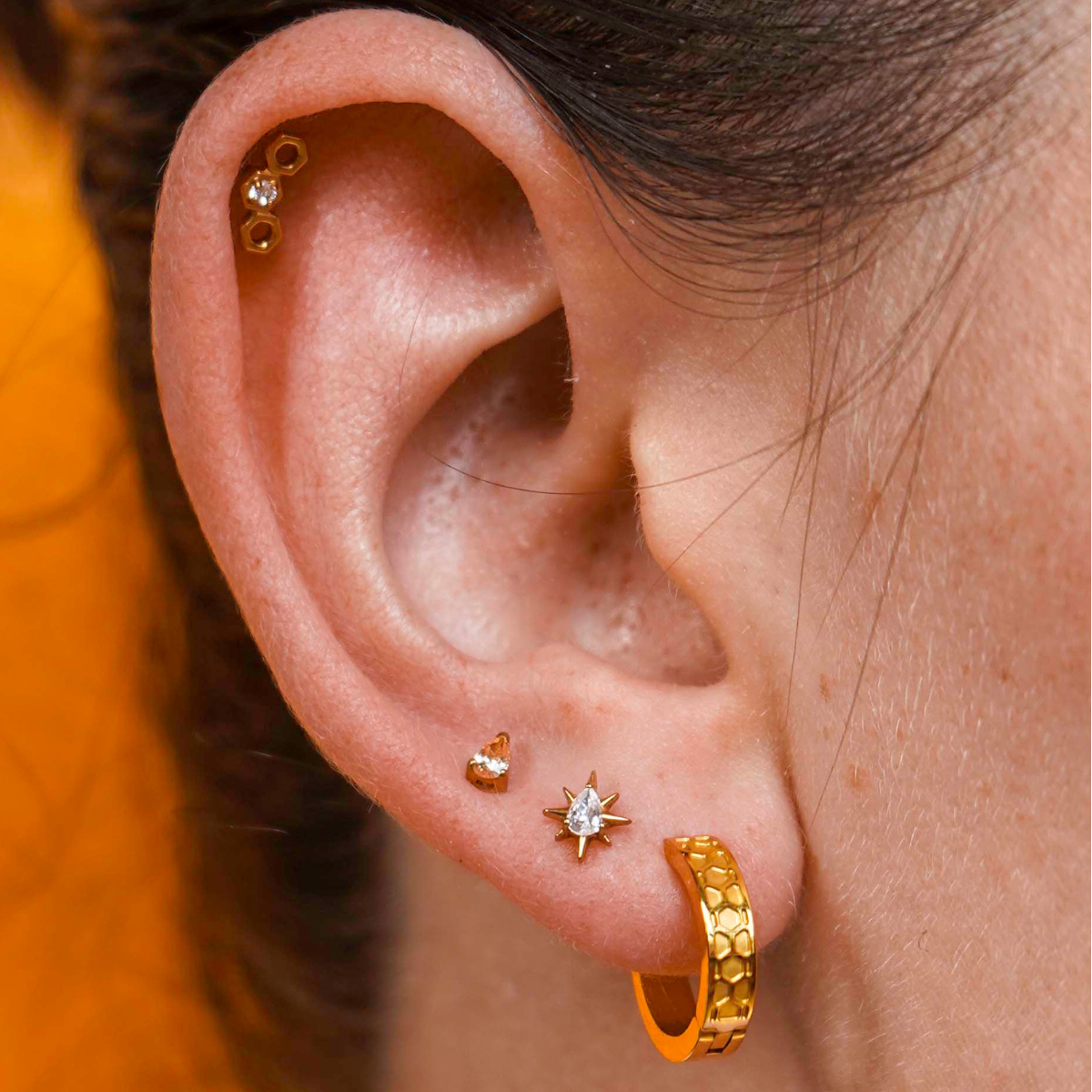 Close up of a model wearing a hexagon shaped flat back stud earring made from 18k gold-plated ASTM F136 implant-grade titanium, with a clear zirconia stone, against a blurred honey-colored background. 