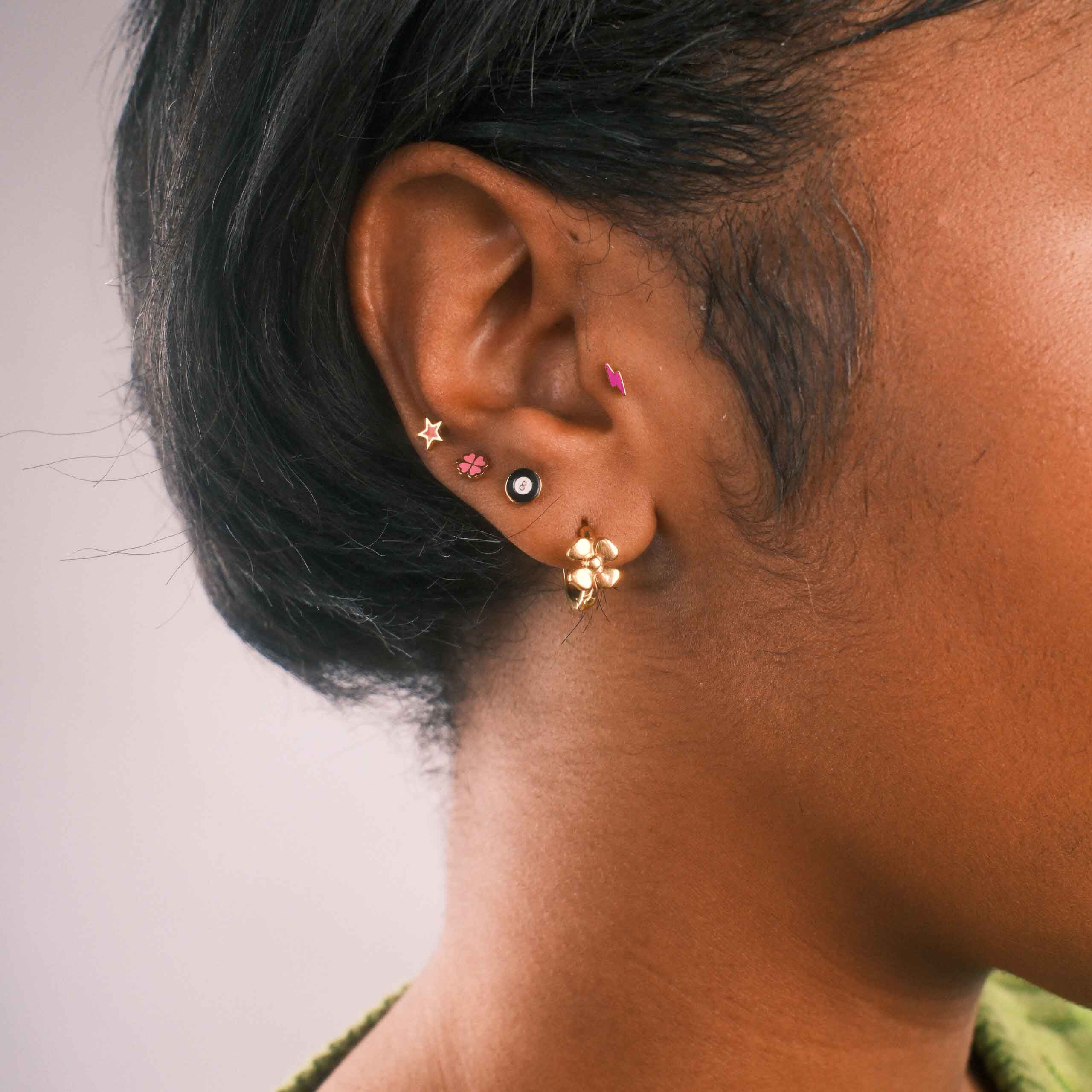 Close-up of an ear with multiple earrings, including a lightning bolt flat back stud in hot pink enamel. 