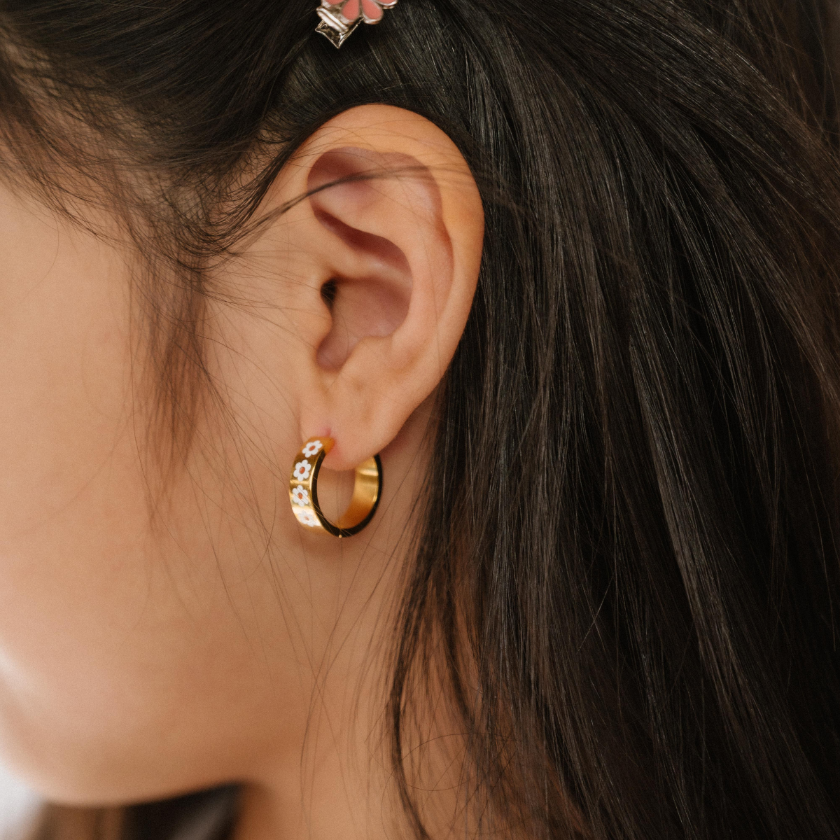 A close up of a model wearing a hoop earring made from 18K gold-plated titanium metal, with a line enamel daisy in white and yellow decorating the hoop.