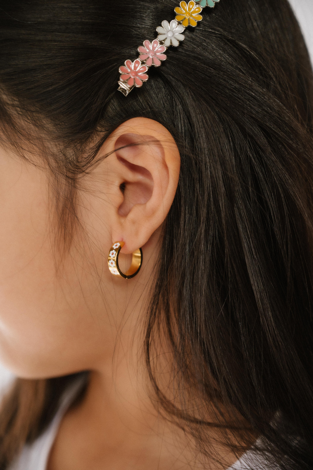 A close up of a model wearing a hoop earring made from 18K gold-plated titanium metal, with a line enamel daisy in white and yellow decorating the hoop.
