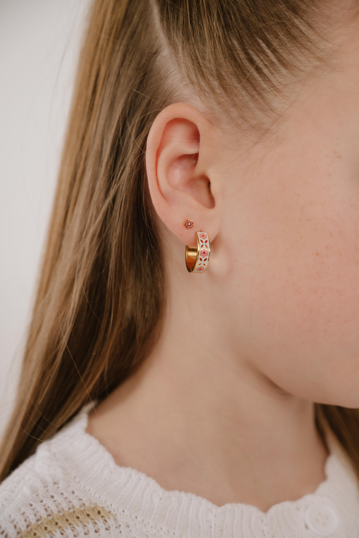 A close up of a model wearing a hoop earring made from gold-plated titanium metal, with folk-art style floral patterns in pink, dark pink, and white enamel.