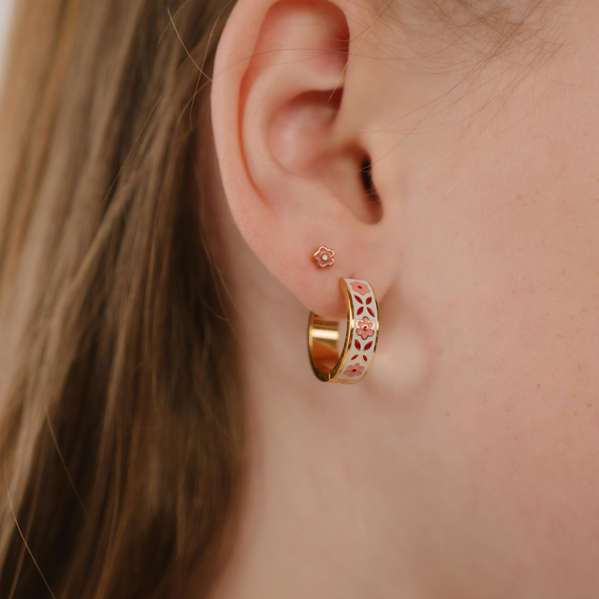 A close up of a model wearing a hoop earring made from 18k gold-plated titanium metal, with folk-art style floral patterns in pink, dark pink, and white enamel.